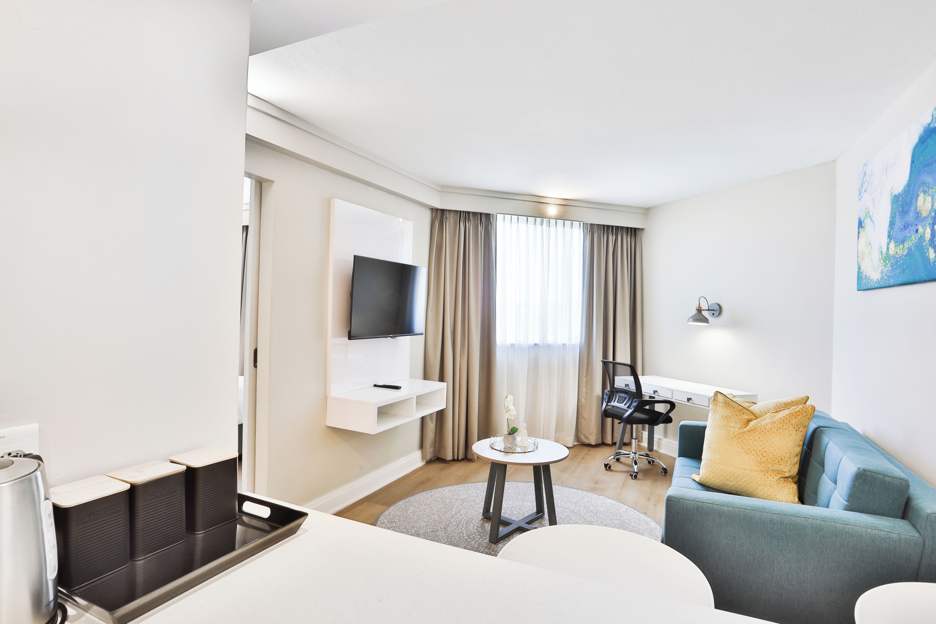 A modern hotel room with a flat-screen TV mounted on the wall, a sofa with cushions, a small round table, a desk with a chair, and a coffee machine.
