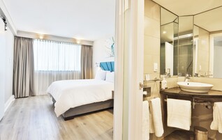 Una habitación de hotel con cama, un ventanal con cortinas, un baño con lavamanos y espejo, y un bote de basura.