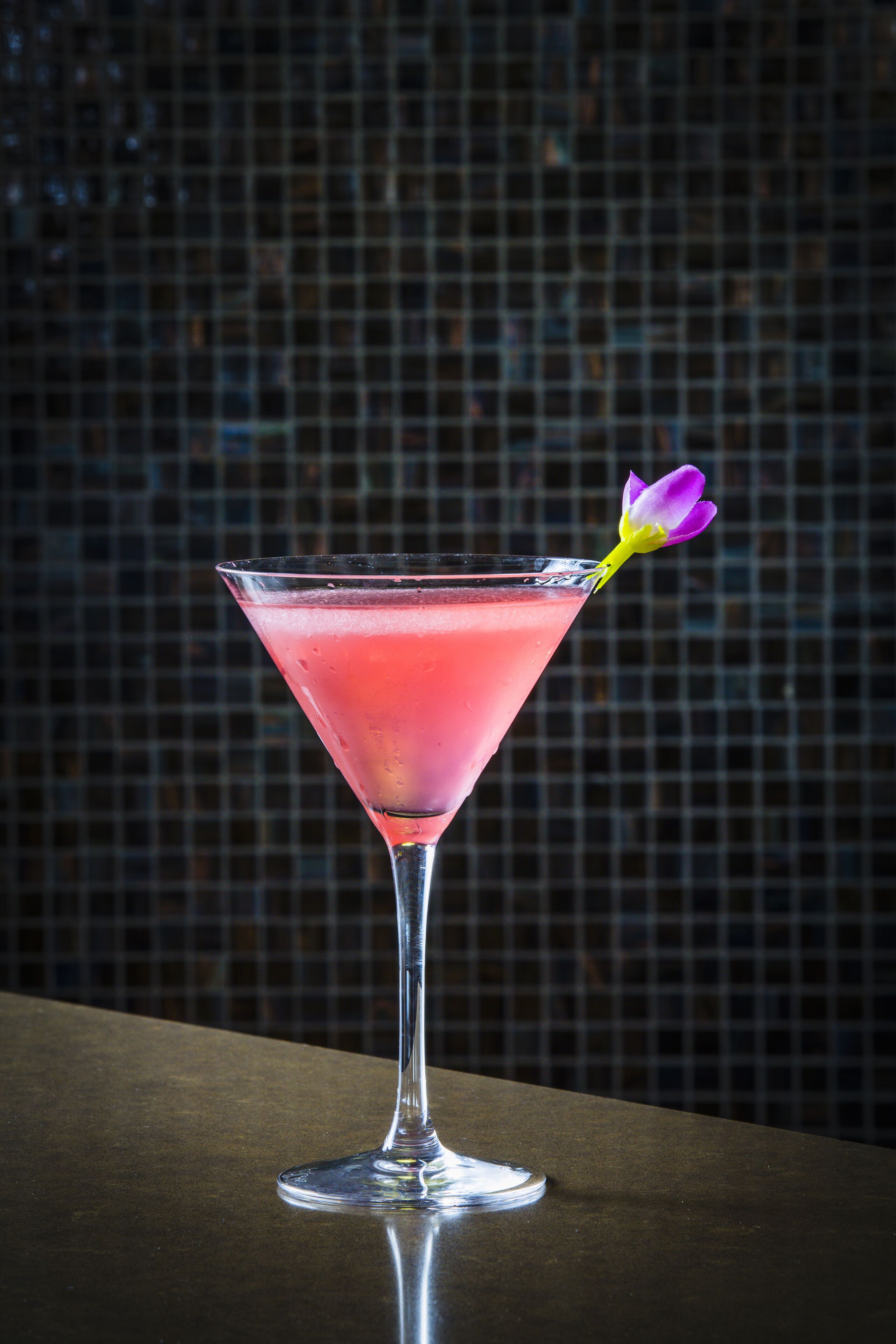 A martini glass with a pink drink and a purple flower garnish on a reflective surface.