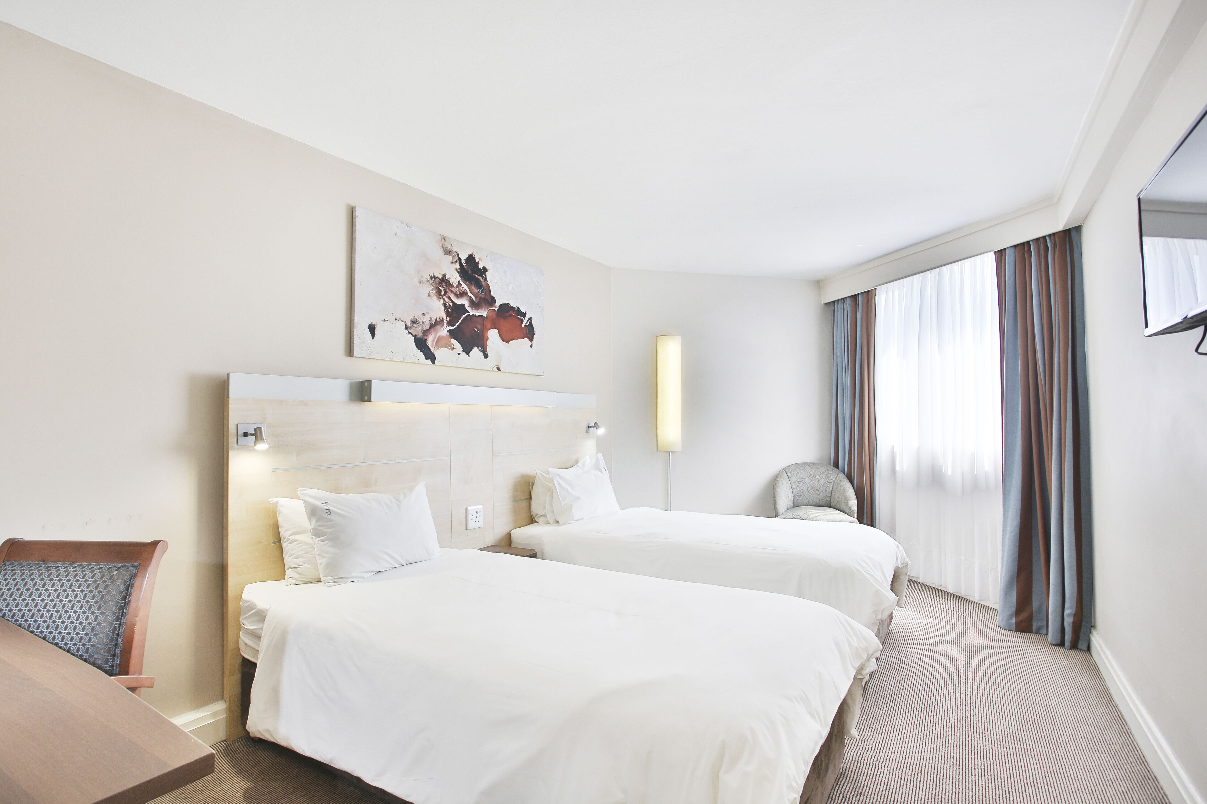 A hotel room with two beds, a desk, a chair, a painting on the wall, and a window with curtains.