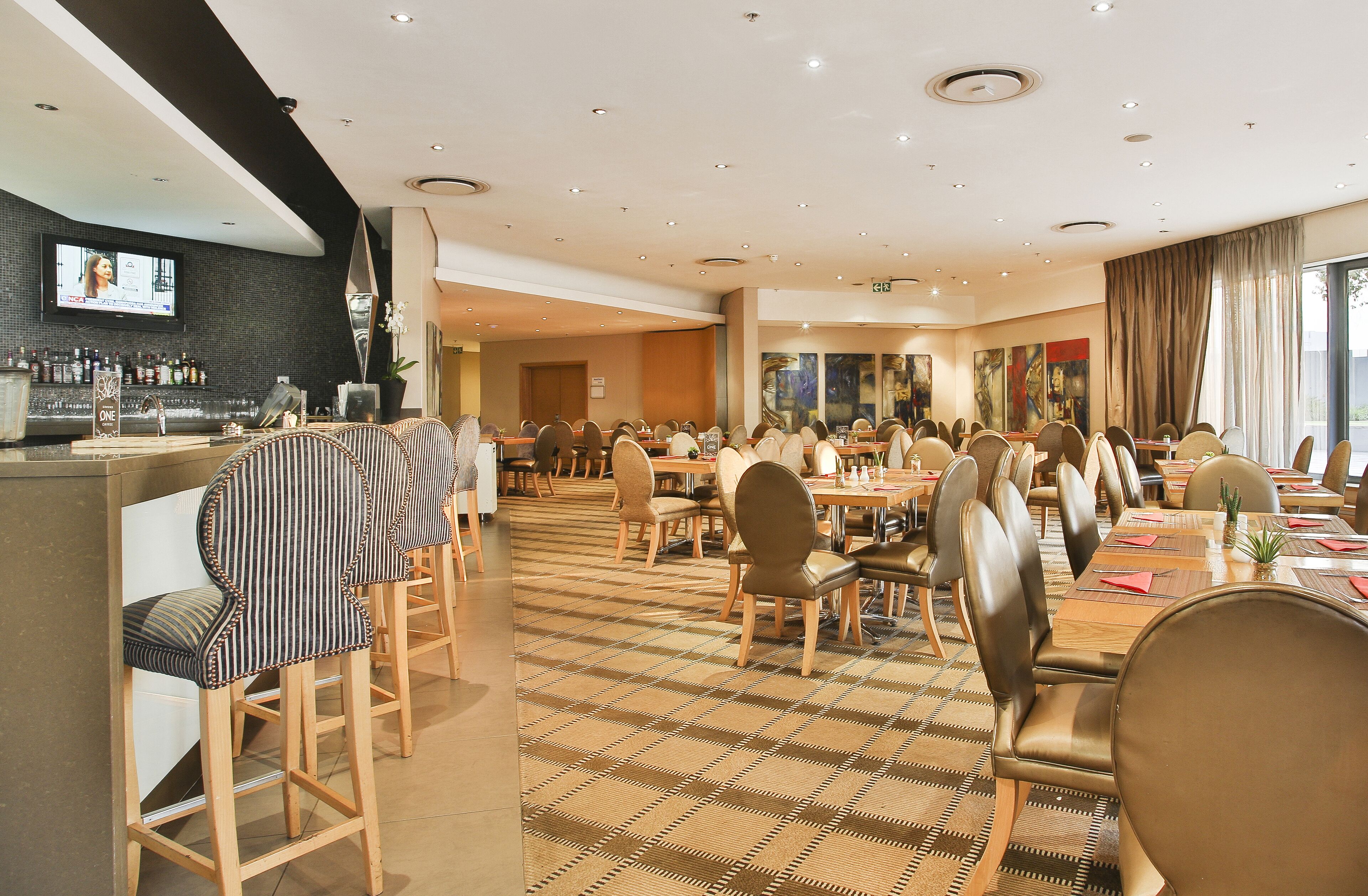 A hotel dining area with multiple tables and chairs, a bar with a television, and artwork on the walls.