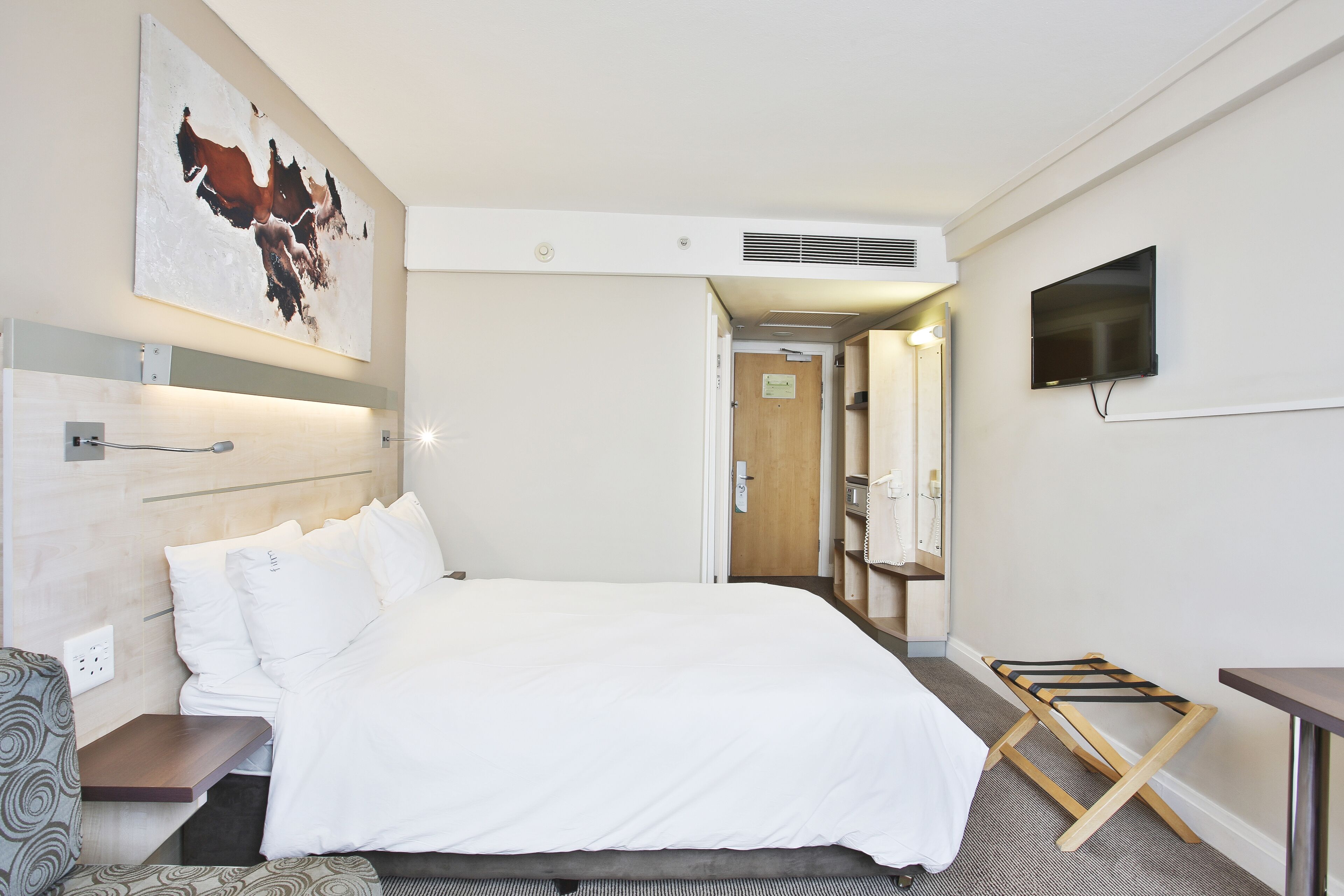 A hotel room with a large bed, a TV mounted on the wall, a wooden side table, and a painting above the bed.