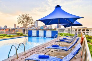 Una piscina en la azotea con sombrillas azules, sillas de descanso y un horizonte urbano al fondo.
