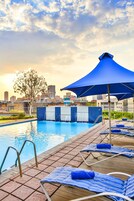 Una piscina con sillas de descanso y sombrillas, y un horizonte urbano al fondo, sobre una terraza con baldosas.