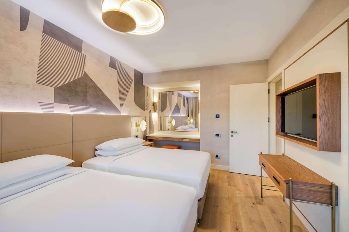 Ein modernes Hotelzimmer mit zwei Betten, einer Holzbank, einem großen Spiegel und einem an der Wand befestigten Fernseher.