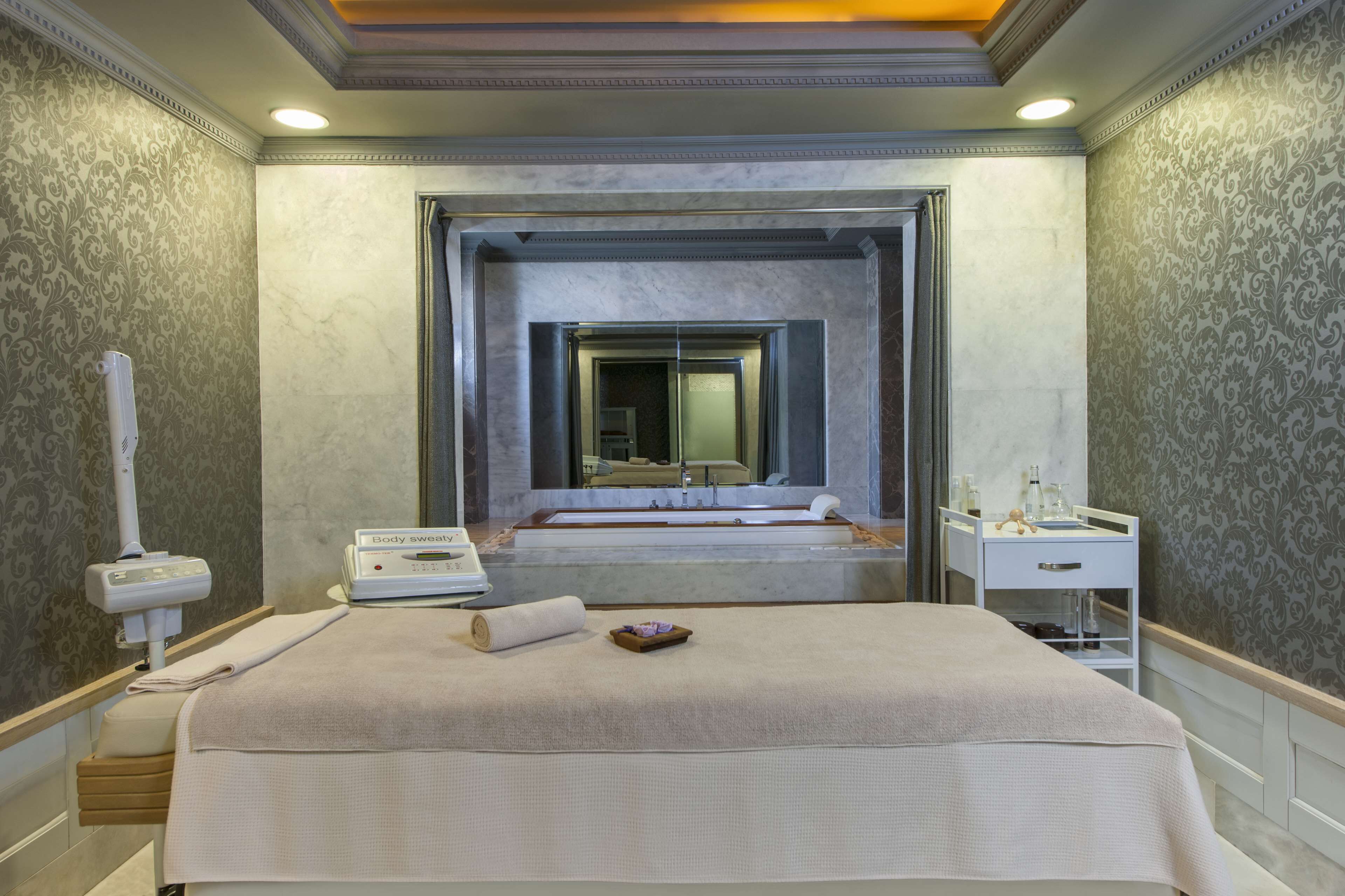 A hotel room with a large bed, a massage chair, a body massage machine, a vanity with a mirror, and a bathtub.