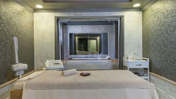 A hotel room with a large bed, a massage chair, a body massage machine, a vanity with a mirror, and a bathtub.