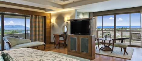 A hotel room with a large bed, a desk with a flat-screen TV, a seating area with a table and chairs, and a balcony with a view of the ocean.