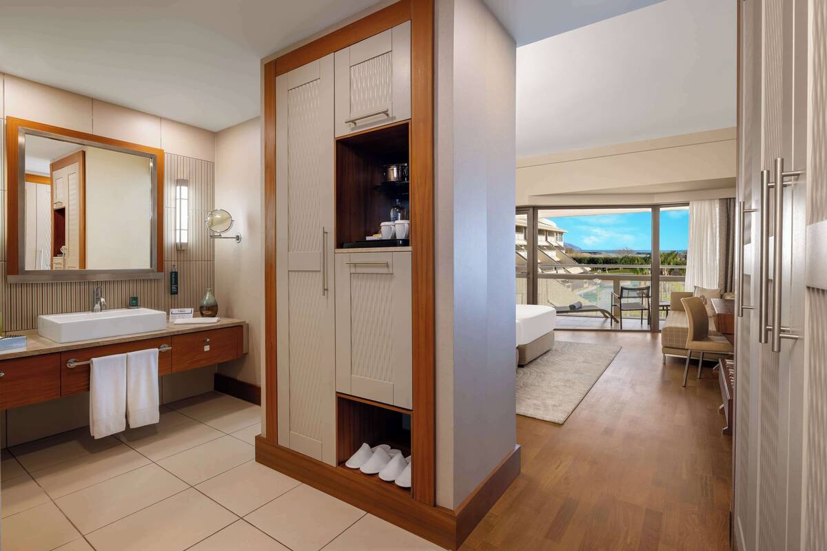 Een moderne badkamer met een grote spiegel, een wastafel en een badkamermeubel. Er is een houten garderobe met een schoenenrek aan de onderkant. De badkamer grenst aan een balkon met uitzicht op de bergen.