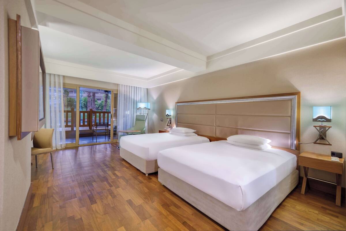 Een hotelkamer met twee bedden, een houten vloer, een groot raam met gordijnen en uitzicht op de omgeving.