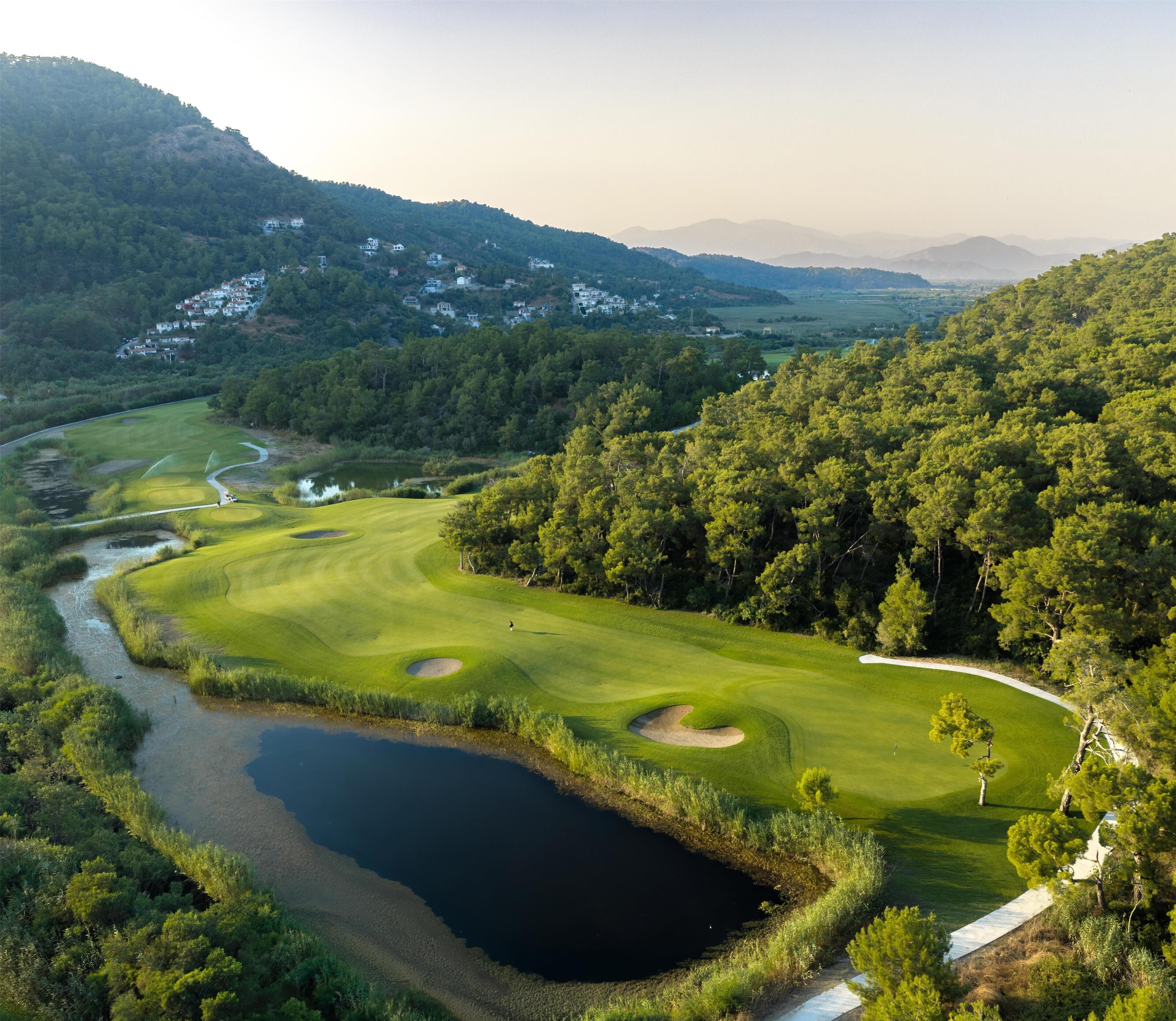 Ein Golfplatz mit Teich, umgeben von dichtem Wald und Hügeln.