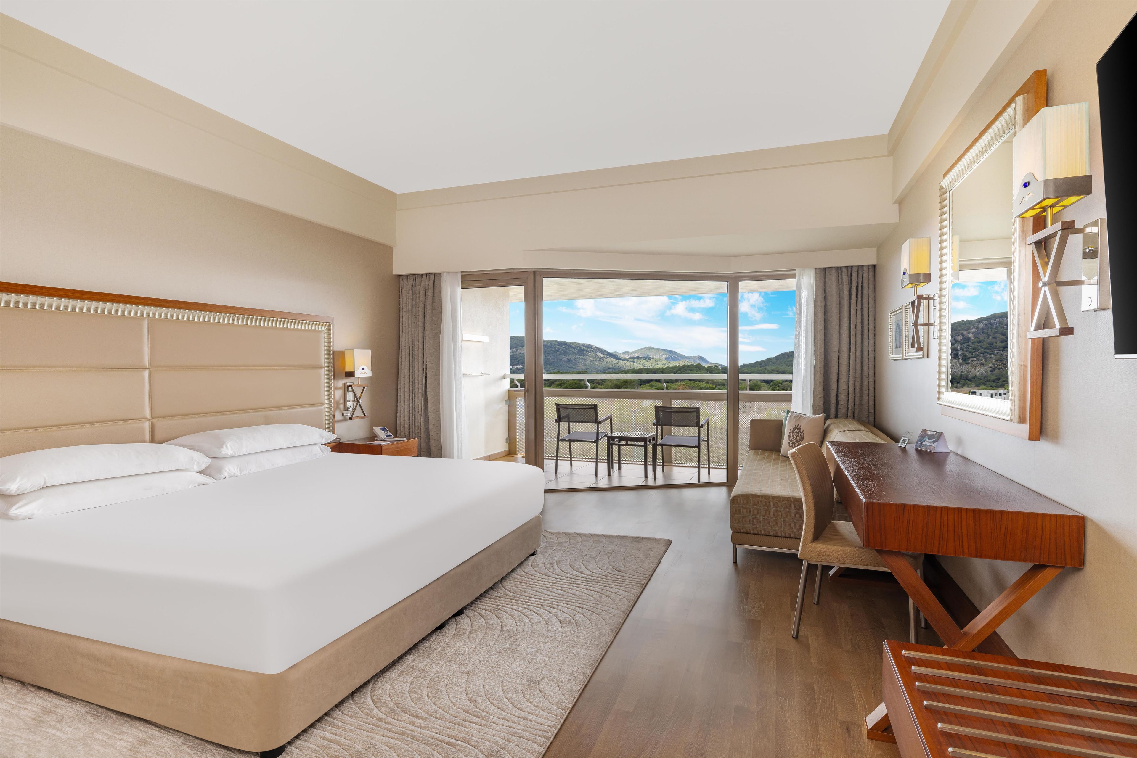 Habitación de hotel con una cama grande, un escritorio y un balcón con vistas a las montañas.