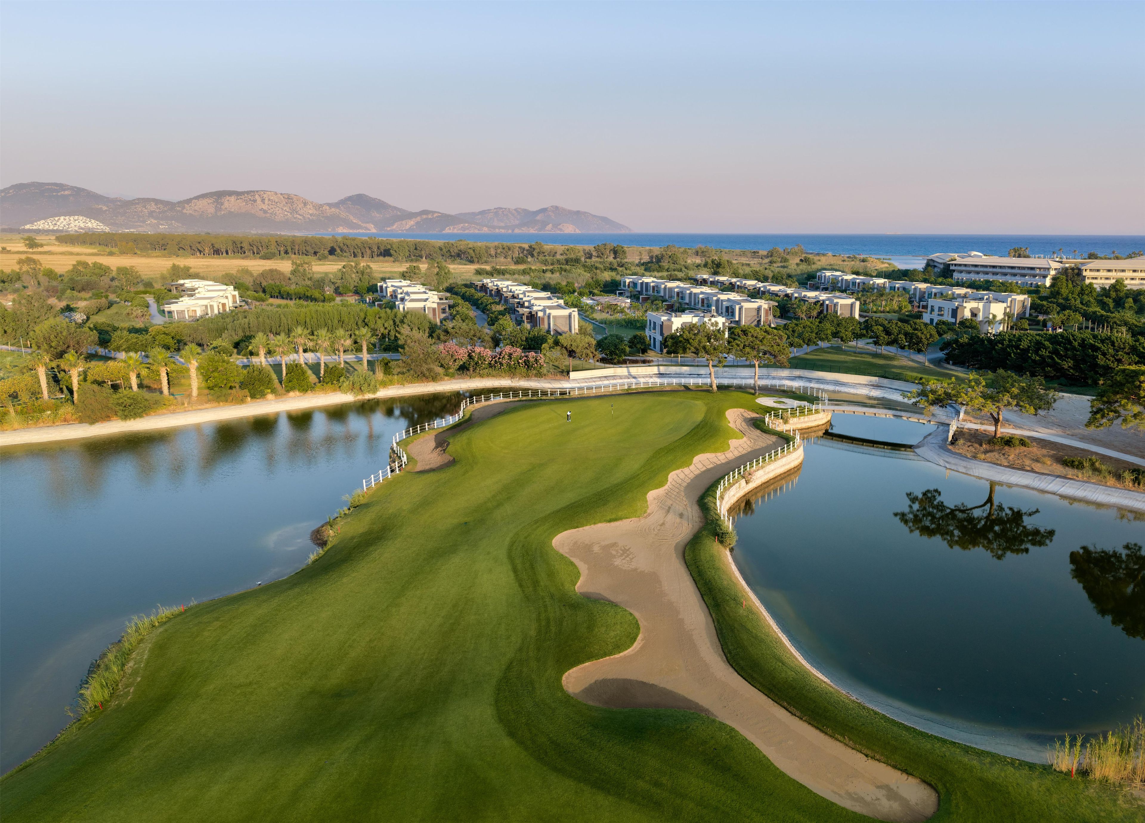 A golf course with a water hazard, surrounded by residential buildings and a mountain range in the background.