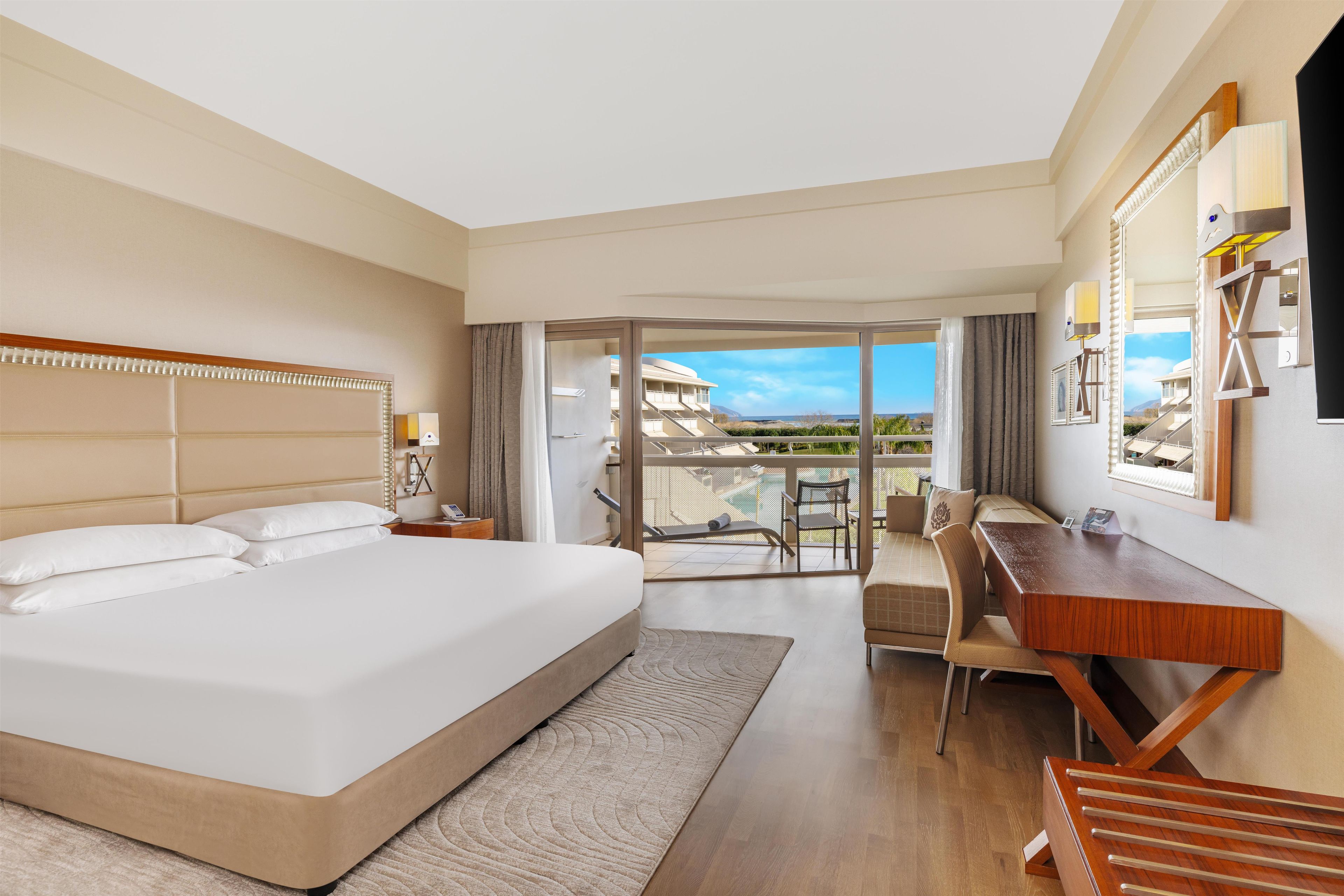 A hotel room with a large bed, a desk, a chair, and a balcony with a view.