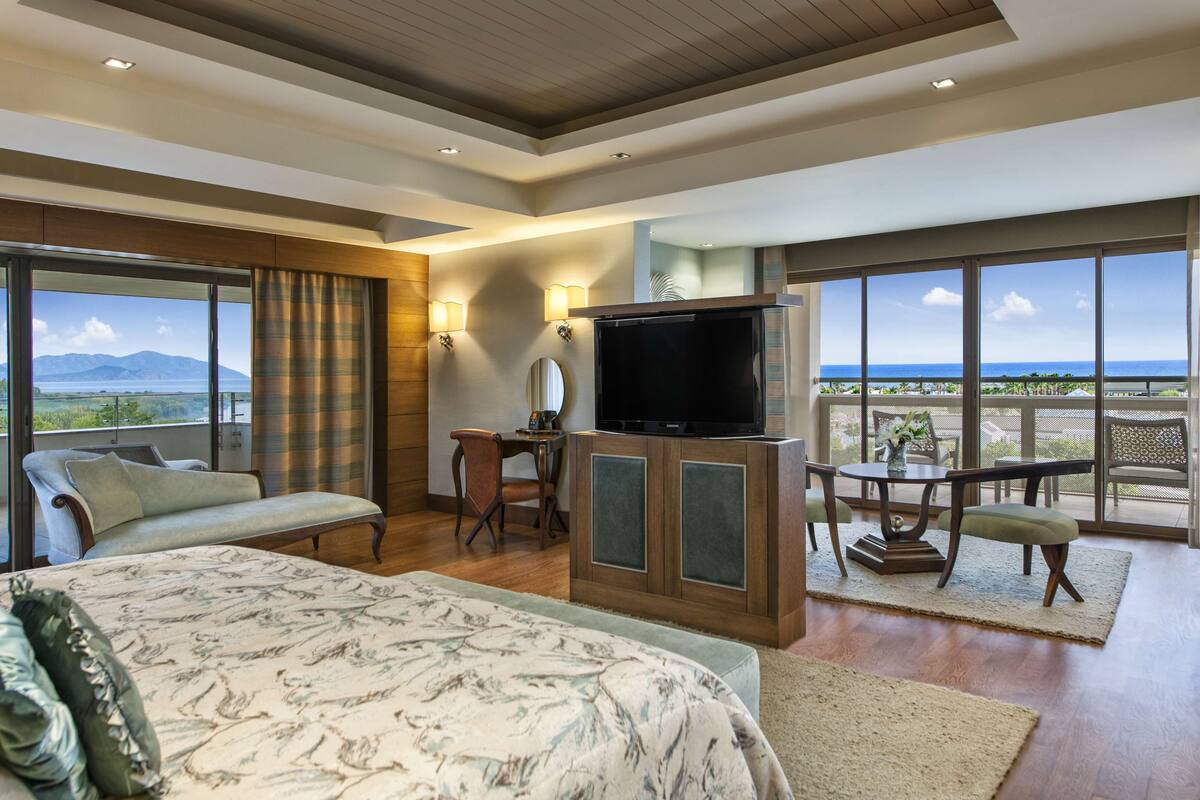 Hotelkamer met een groot bed, een bureau met een televisie, een eethoek en grote ramen met uitzicht op de oceaan.