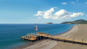 Eine Holzstege ragt ins Meer, mit Liegestühlen und Flaggen, ein Kiesstrand und ein Berg im Hintergrund.