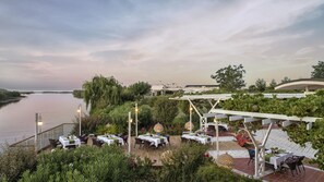 Un espace de restauration en plein air avec des tables dressées pour les clients, offrant une vue sur un plan d’eau et un jardin aménagé.