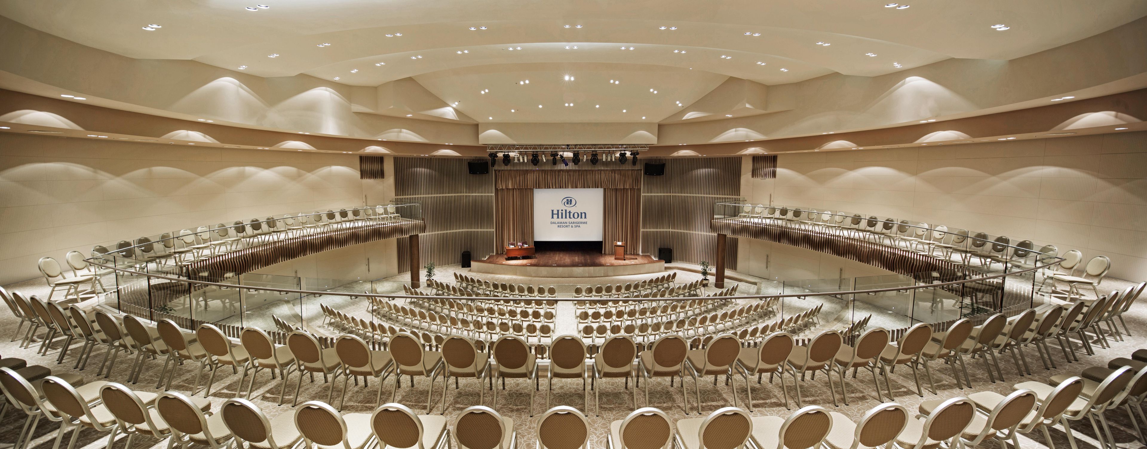 Un salón de conferencias con un escenario, asientos dispuestos en forma de semicírculo y una pantalla grande que muestra el logo de Hilton.