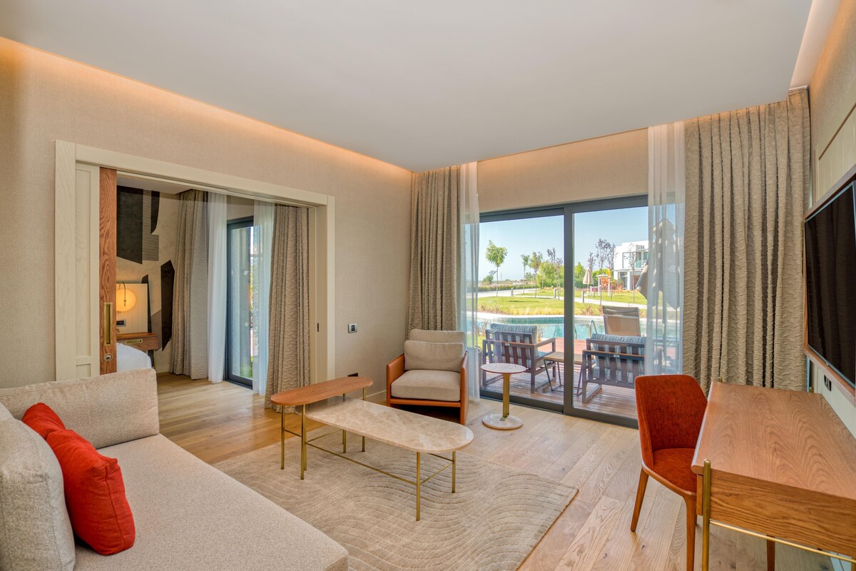 Een moderne hotelkamer met een bank, fauteuil en een eettafel. Er is een groot raam met gordijnen, een flatscreen-tv en uitzicht op een zwembad en groen.