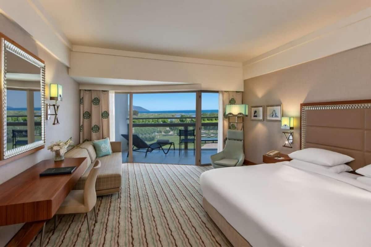 Ein Hotelzimmer mit einem großen Bett, einem Schreibtisch, einem Stuhl, einem Balkon mit Aussicht und einem Nachttisch mit Lampe.