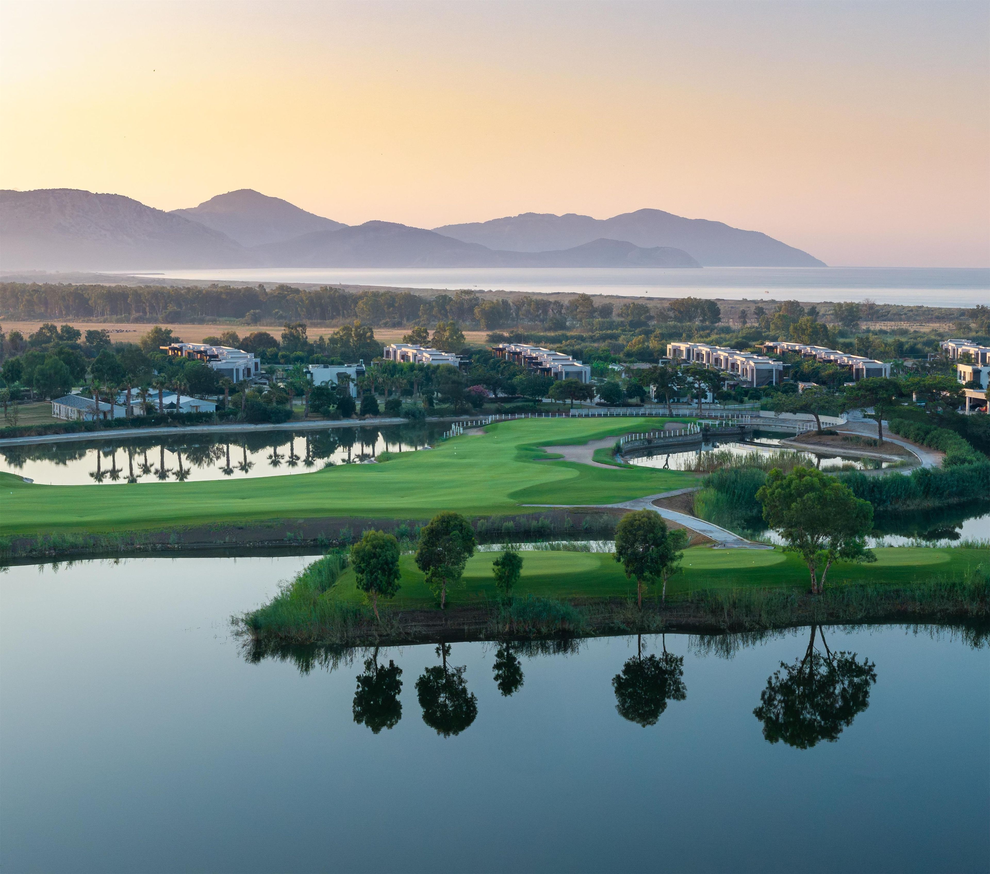 A golf course with a lake in the foreground, surrounded by buildings and trees.