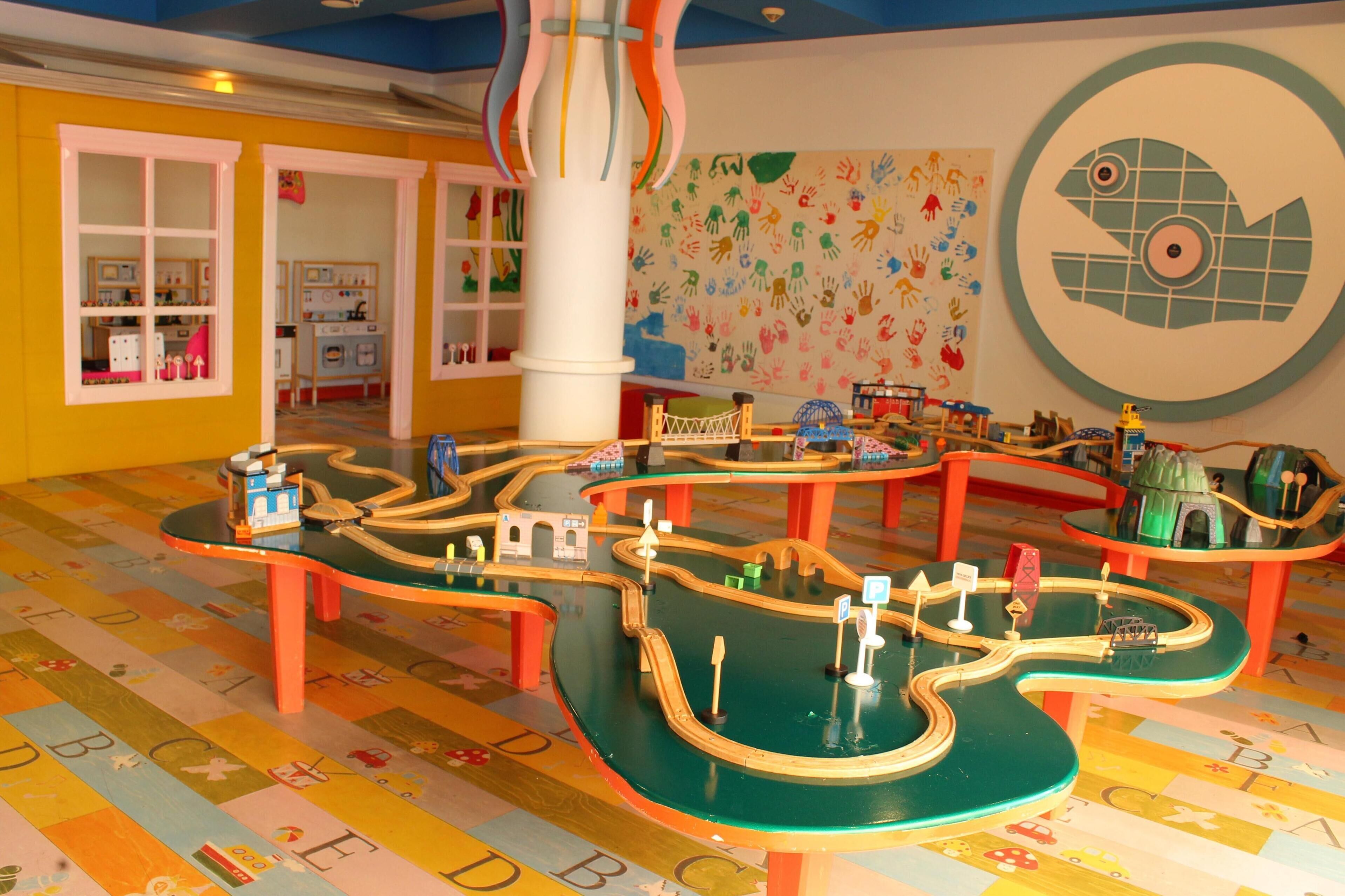 Zona infantil con suelo tipo laberinto, estructuras de juego coloridas y una pared con huellas de manos.