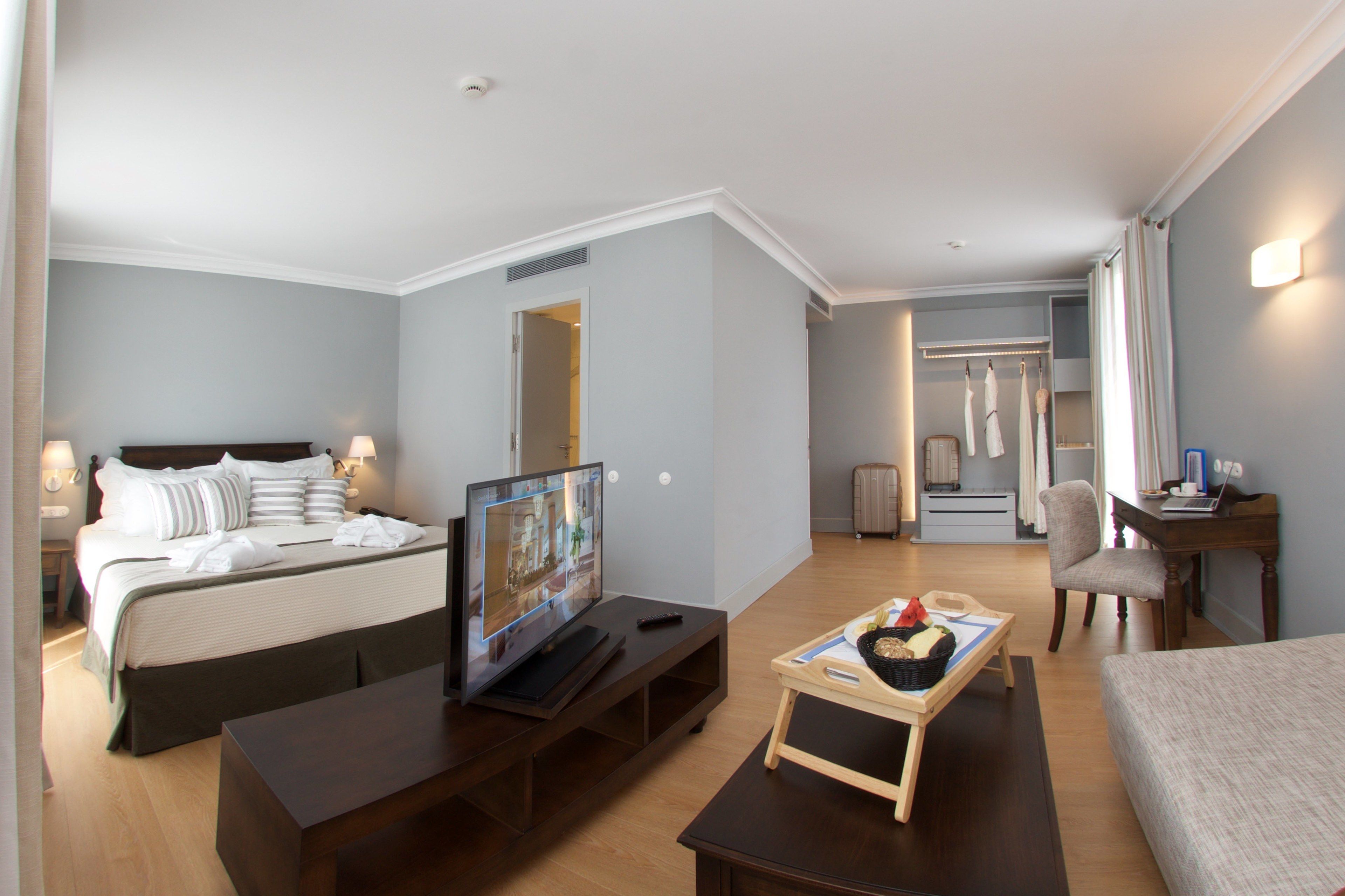 A hotel room with a bed, TV, bedside table, wooden bench, and a desk with a laptop.