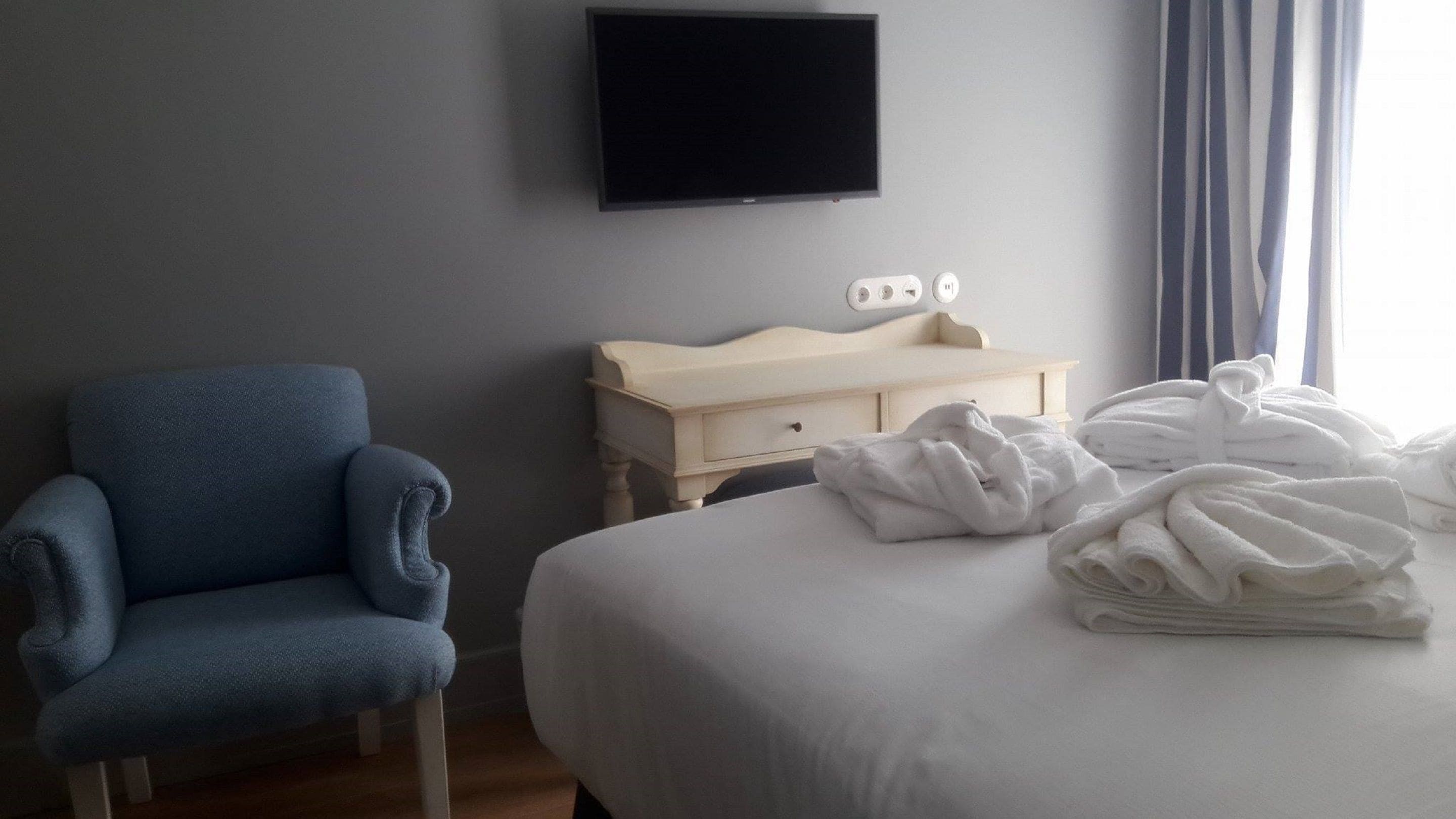 Een hotelkamer met een bed, een blauwe fauteuil, een nachtkastje met een lamp en een aan de muur bevestigde televisie.