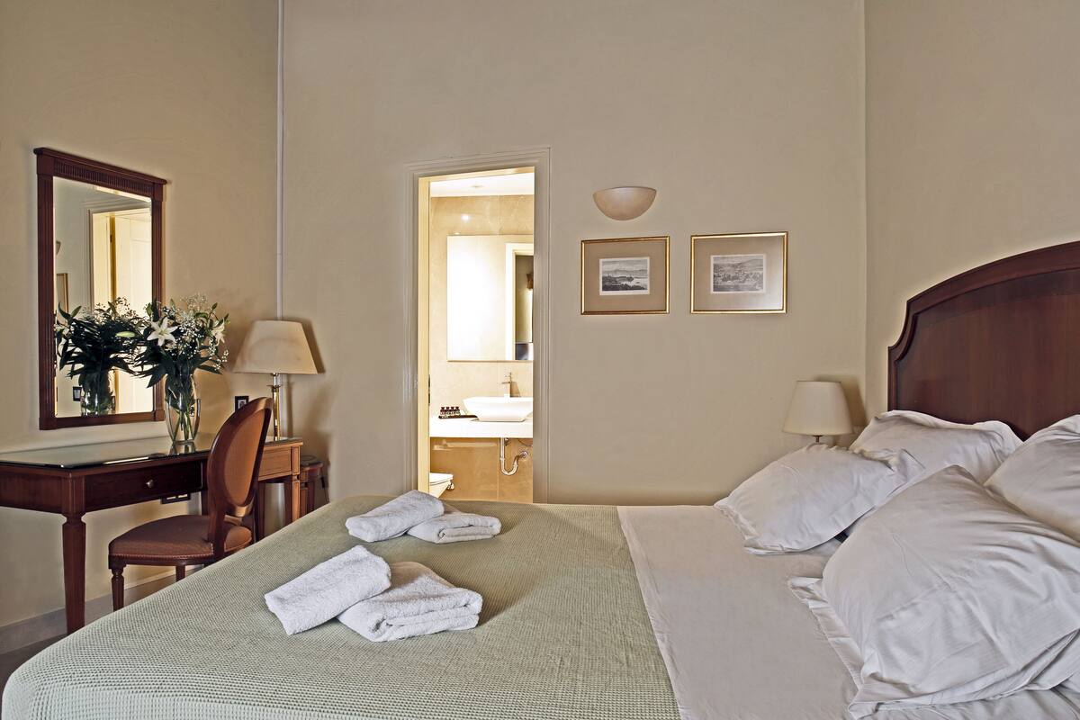 Camera d'albergo con un letto, una scrivania con una sedia, uno specchio e un bagno visibile attraverso una porta aperta.