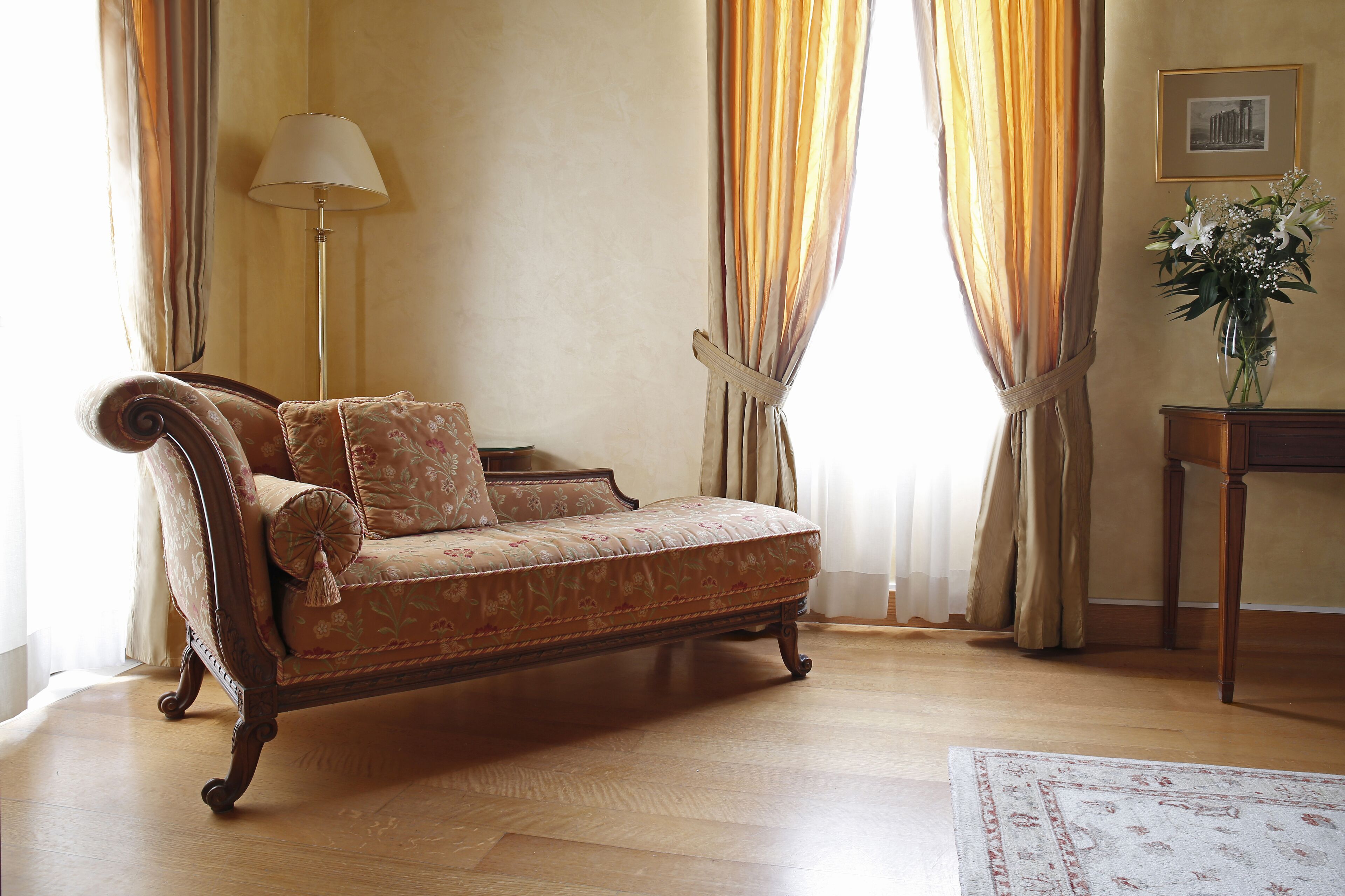 A vintage sofa with floral upholstery in a room with wooden flooring, a standing lamp, and a vase with flowers on a side table.