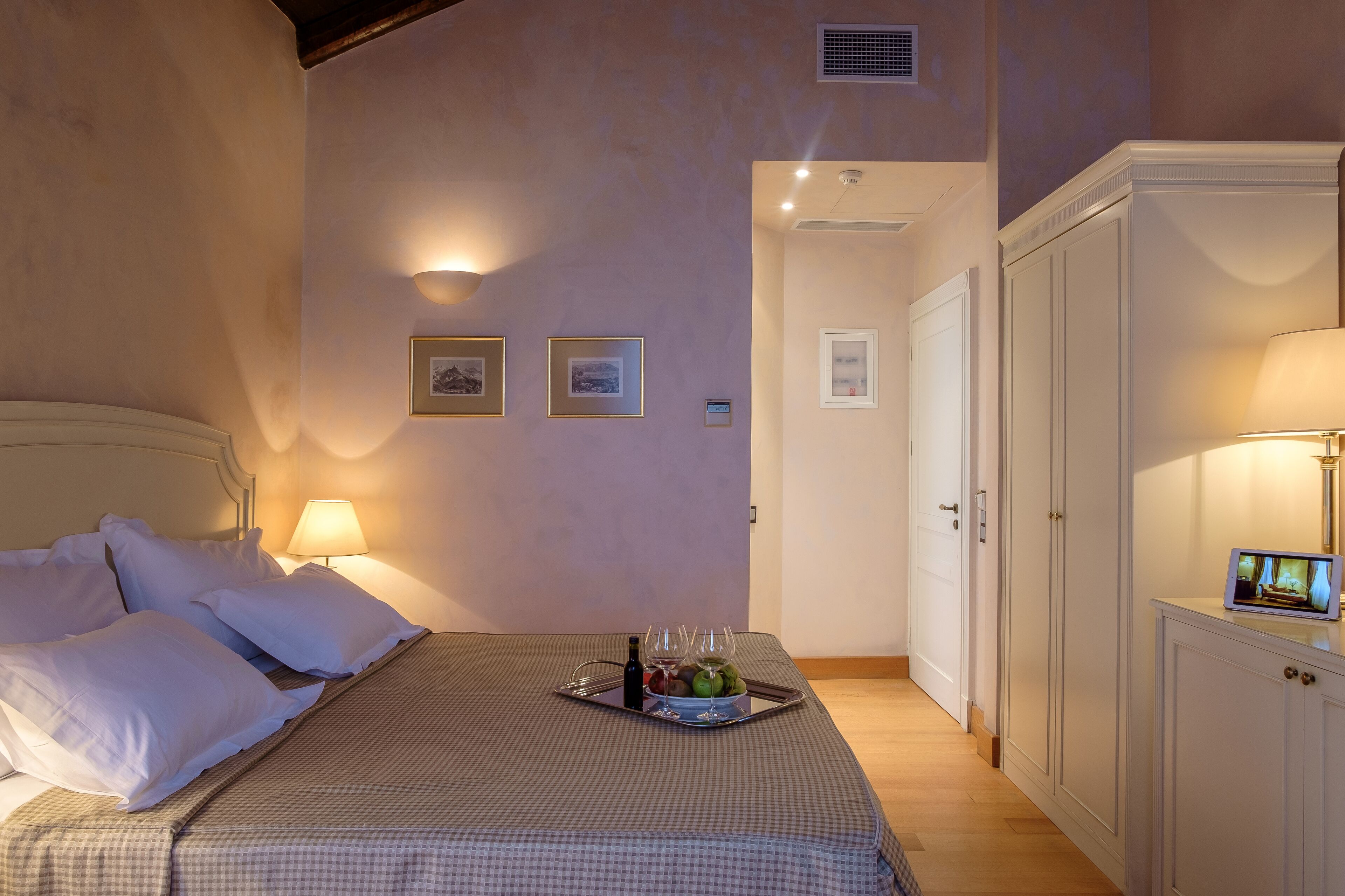 Une chambre à coucher avec un lit, des oreillers, un plateau avec des verres à vin et des fruits, une lampe de chevet et un téléviseur posé sur une commode.