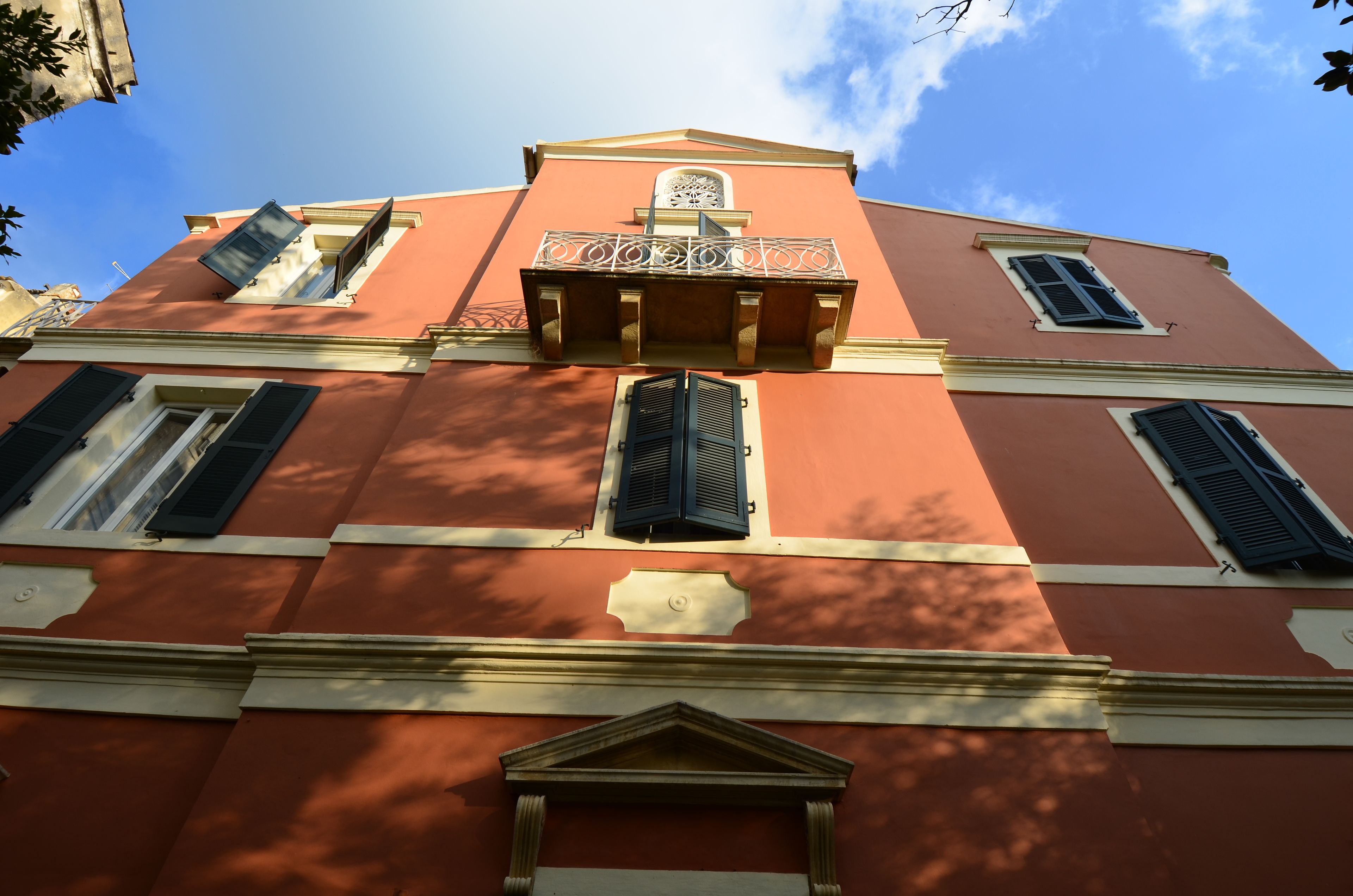 Un edificio con fachada roja, varias ventanas con contraventanas y un balcón con barandilla decorativa.