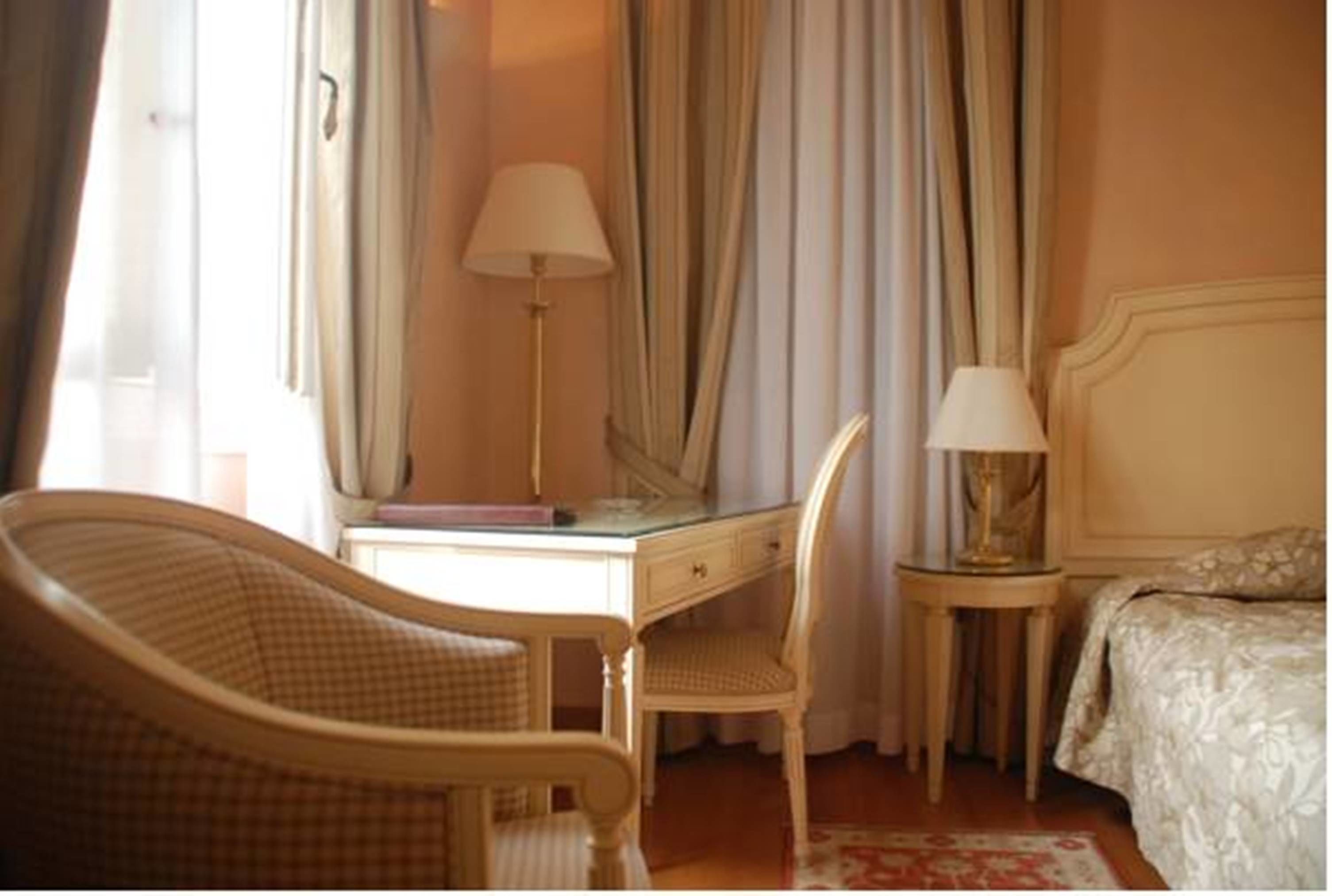 Une chambre d’hôtel avec un lit, un bureau et une chaise, une lampe et une fenêtre avec des rideaux.