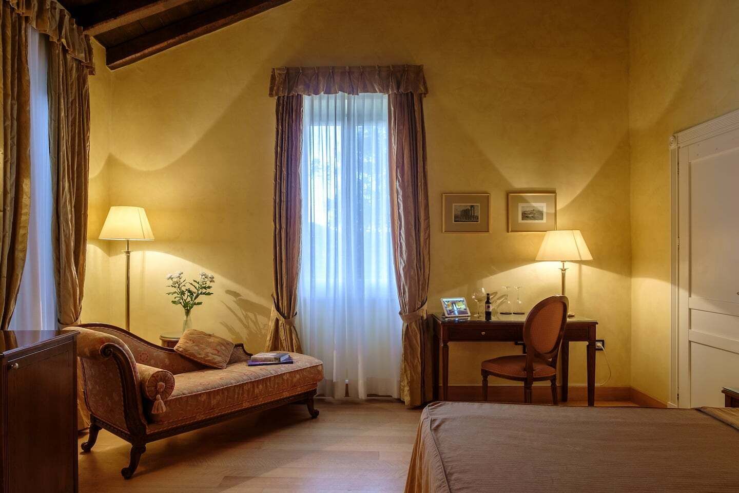 Habitació d'hotel amb llit, escriptori, cadira, làmpada i finestra amb cortines.
