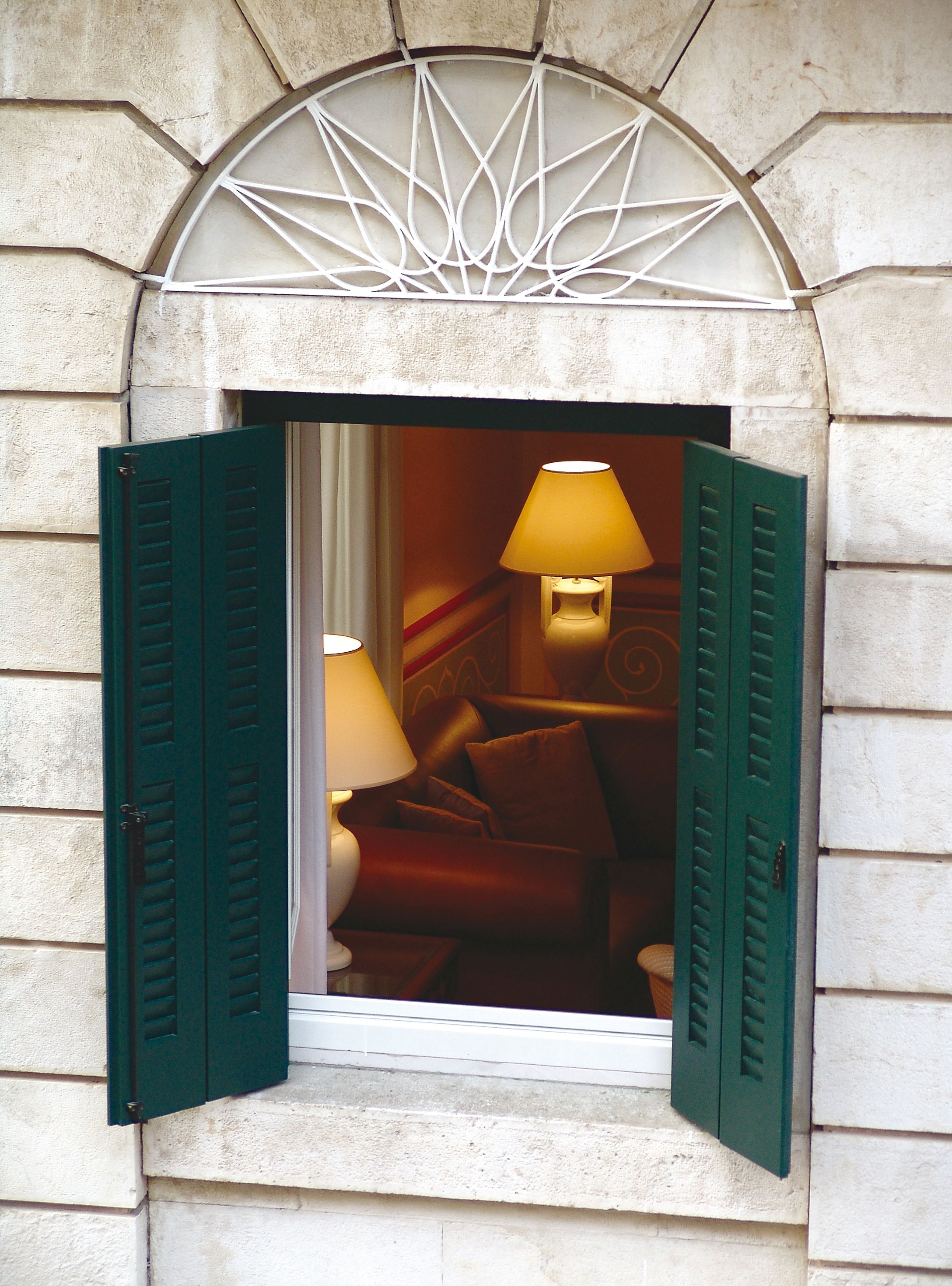 A window with green shutters, a lamp, and a glimpse of a room interior.