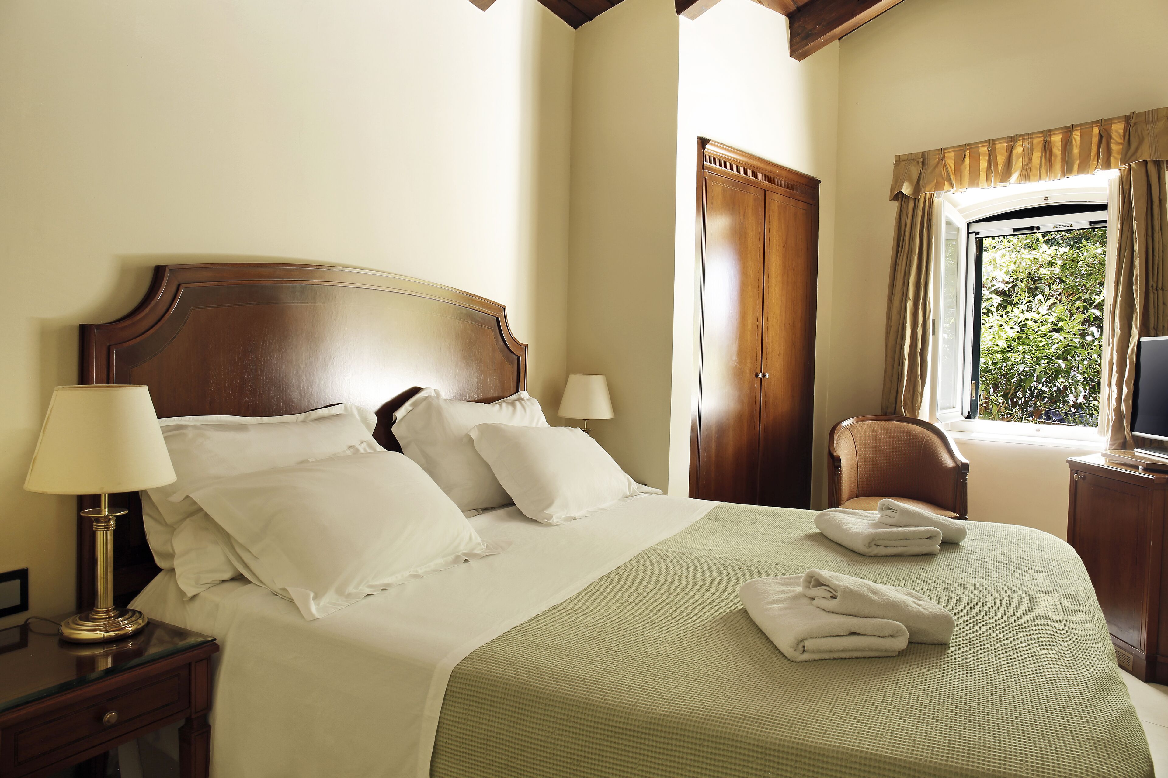 A neatly made bed with pillows and a green bedspread, a bedside lamp, a chair, and a television on a wooden nightstand.