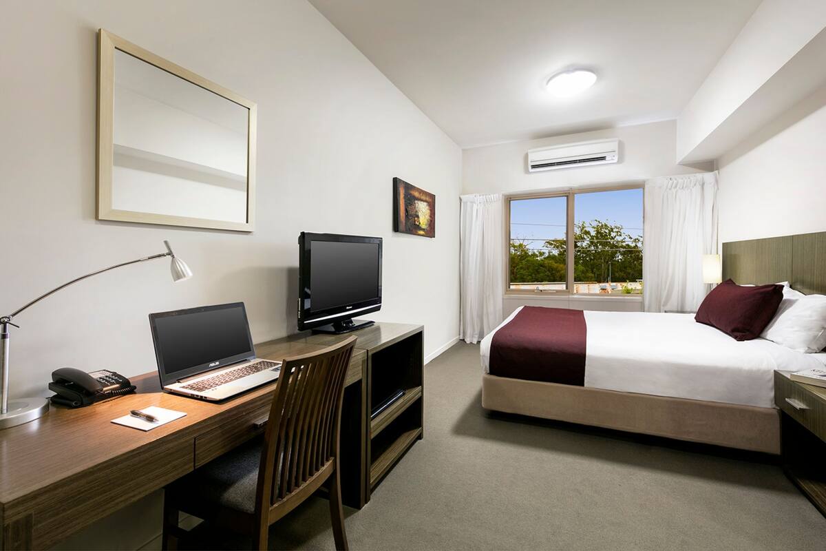 Habitació d'hotel amb llit, escriptori, ordinador portàtil, televisió i finestra amb cortines.