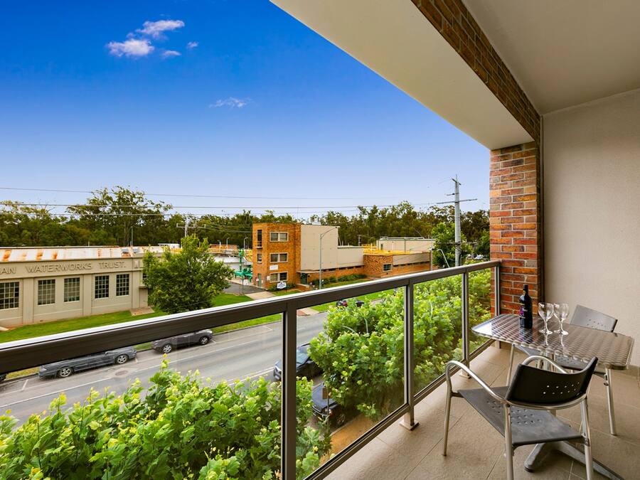 Varanda com mesa e cadeiras, vista para uma rua com edifícios e áreas verdes.