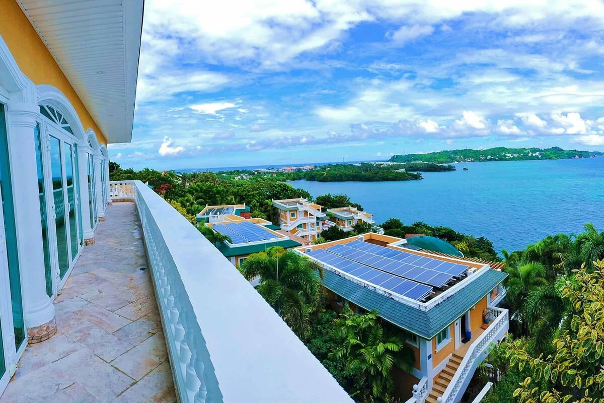 Un balcón con vistas a la costa, paneles solares en un edificio y abundante vegetación.