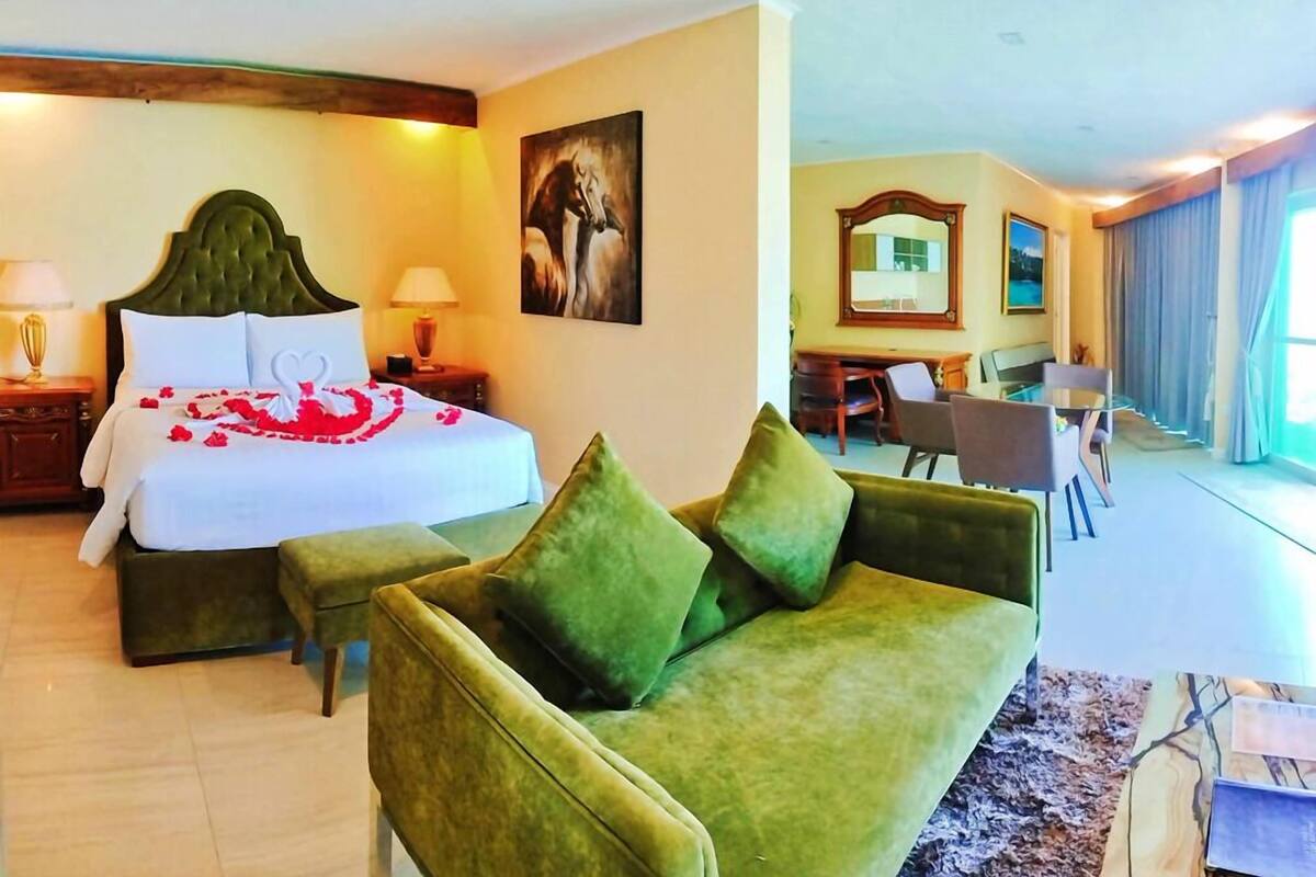 Habitación de hotel con cama, sofá verde y zona de comedor con mesa y sillas.
