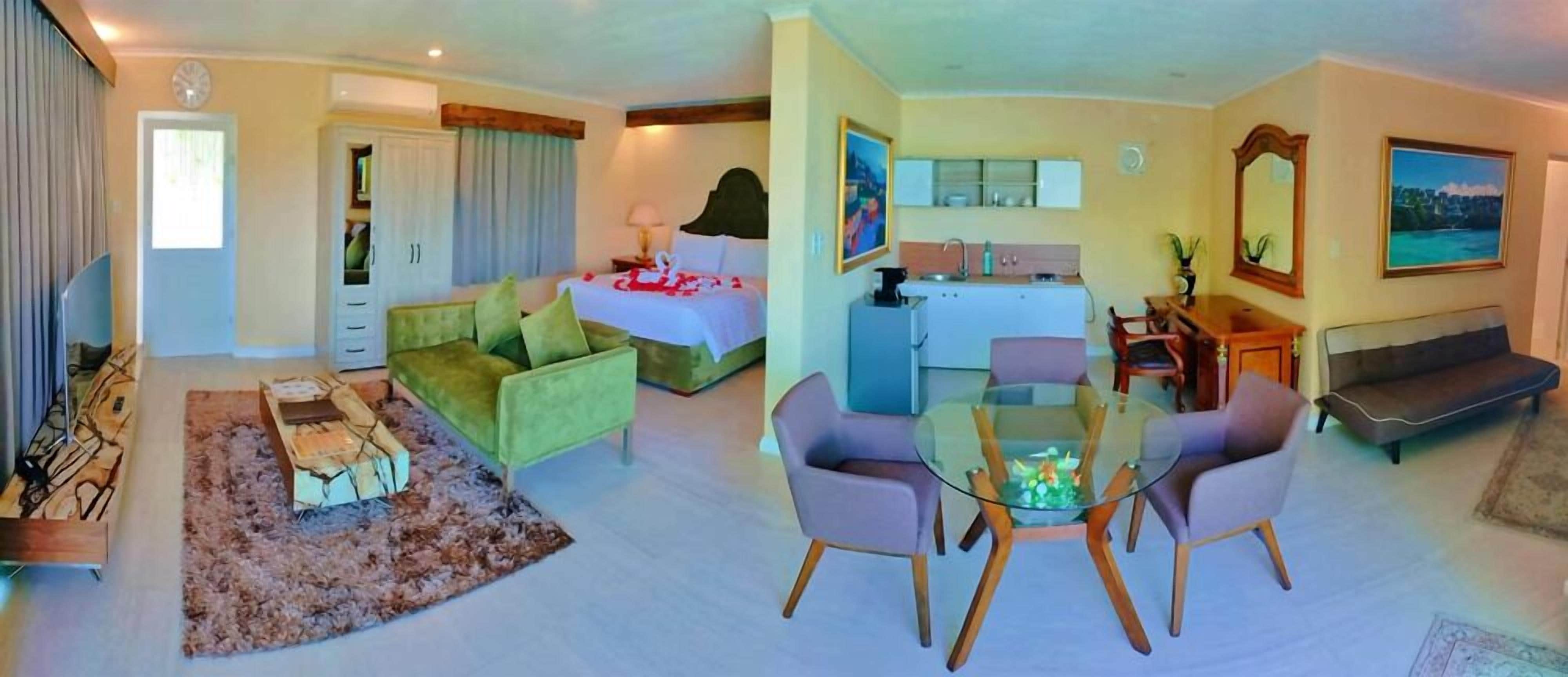 A hotel room with a bed, a sofa, a dining table with chairs, a kitchen area, and a living room with a TV.
