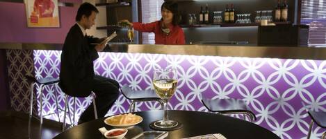 A bar area with a purple patterned wall, a counter with bottles and glasses, and a table with a wine glass, bread, and a menu.