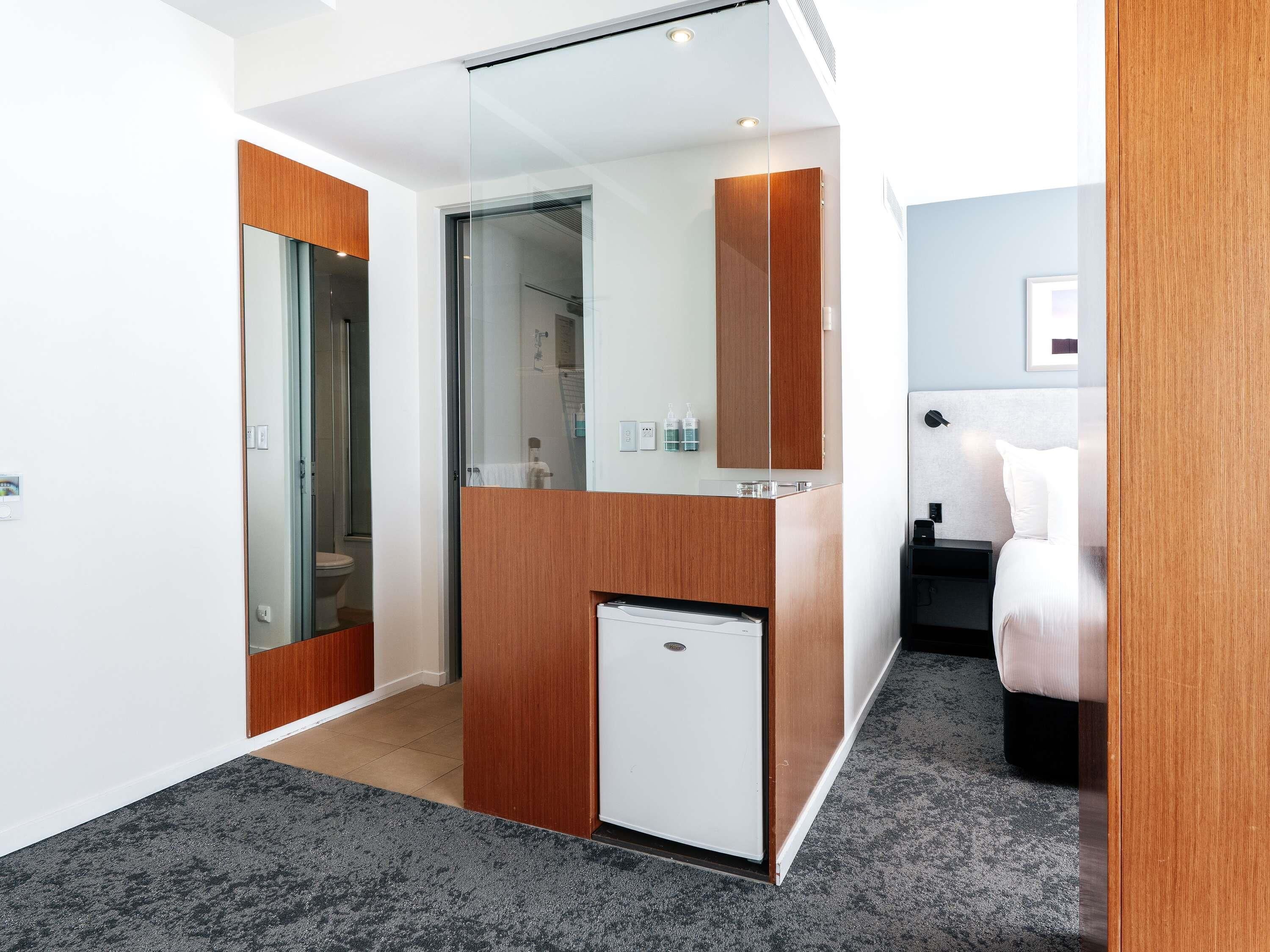 A modern hotel room with a compact layout, including a bed, a small kitchenette, and a glass partition separating the bathroom.