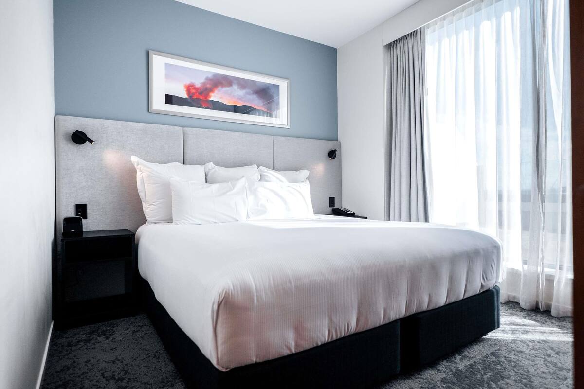 Ein modernes Hotelzimmer mit einem großen Bett, einem Kopfteil mit einem gerahmten Bild, einem Nachttisch mit Telefon und einem Fenster mit leichtem Vorhang.