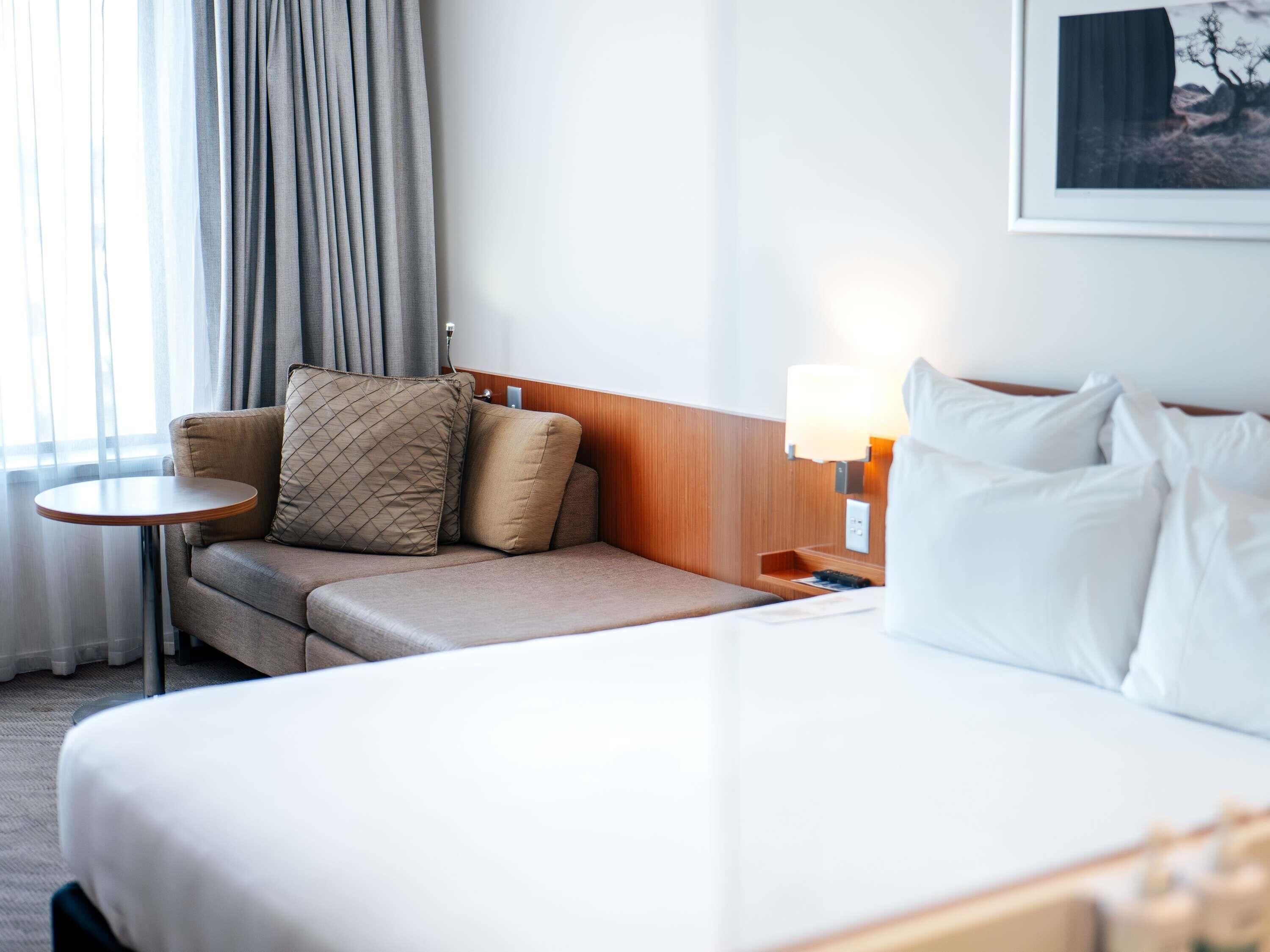 A hotel room with a bed, a small round table, a sofa with cushions, a lamp, and a framed picture on the wall.