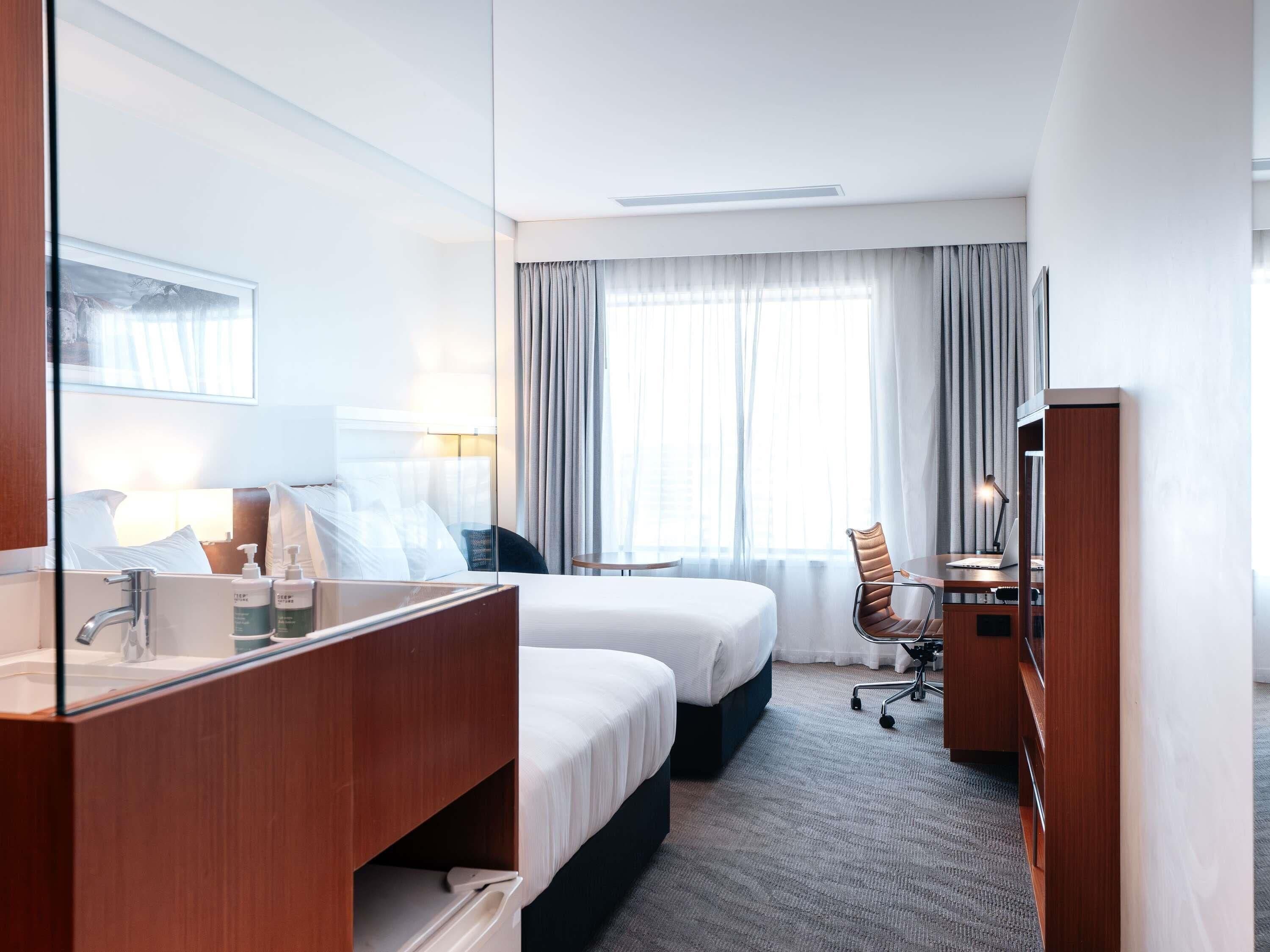 A hotel room with a large mirror, two beds, a desk with a chair, and a window with curtains.