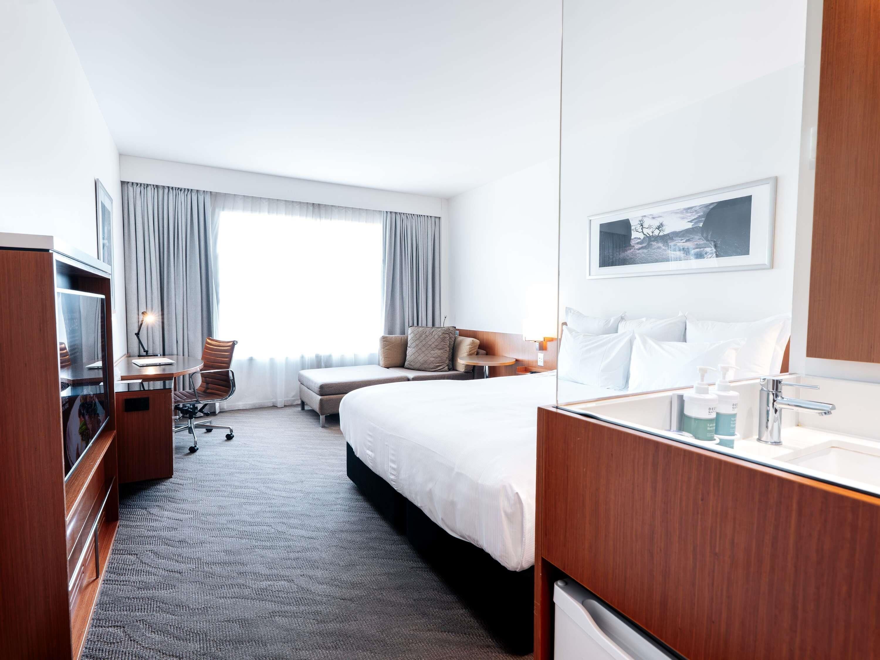 Ein Hotelzimmer mit Bett, Schreibtisch, Stuhl, Fernseher und einem Badezimmer mit Waschbecken und Pflegeprodukten. Ein großes Fenster mit Vorhängen rundet das Zimmer ab.