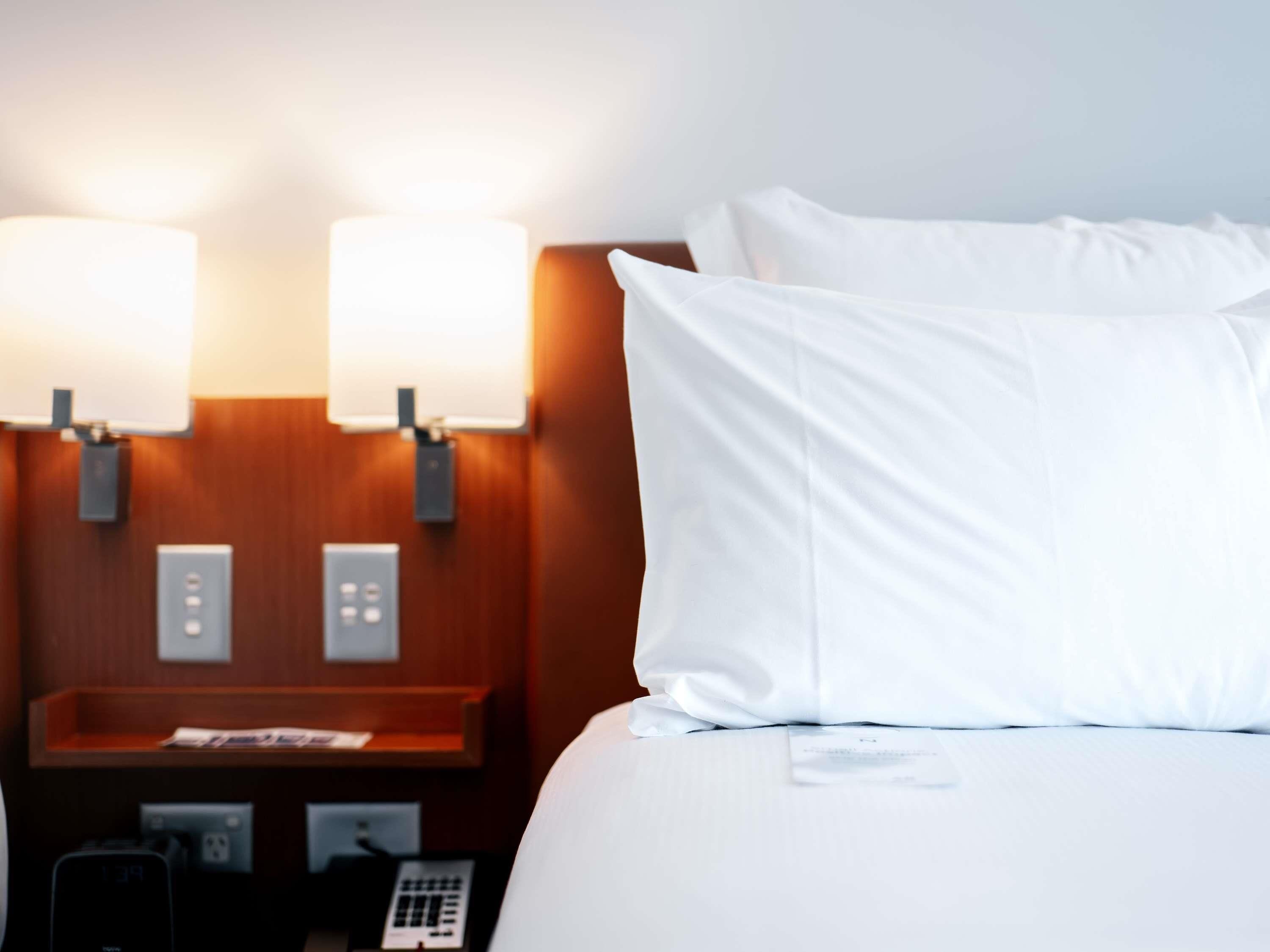Ein Hotelzimmer mit einem ordentlich bezogenen Bett, zwei Wandleuchten, einem Nachttisch aus Holz mit Telefon und einem kleinen Tisch sowie einer Steckdose.
