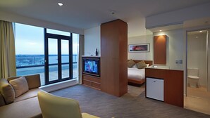 A modern hotel room with a large window, a sofa, a chair, a bed, a TV, a mini-fridge, and a bathroom. - Novotel Christchurch Cathedral Square (Christchurch)