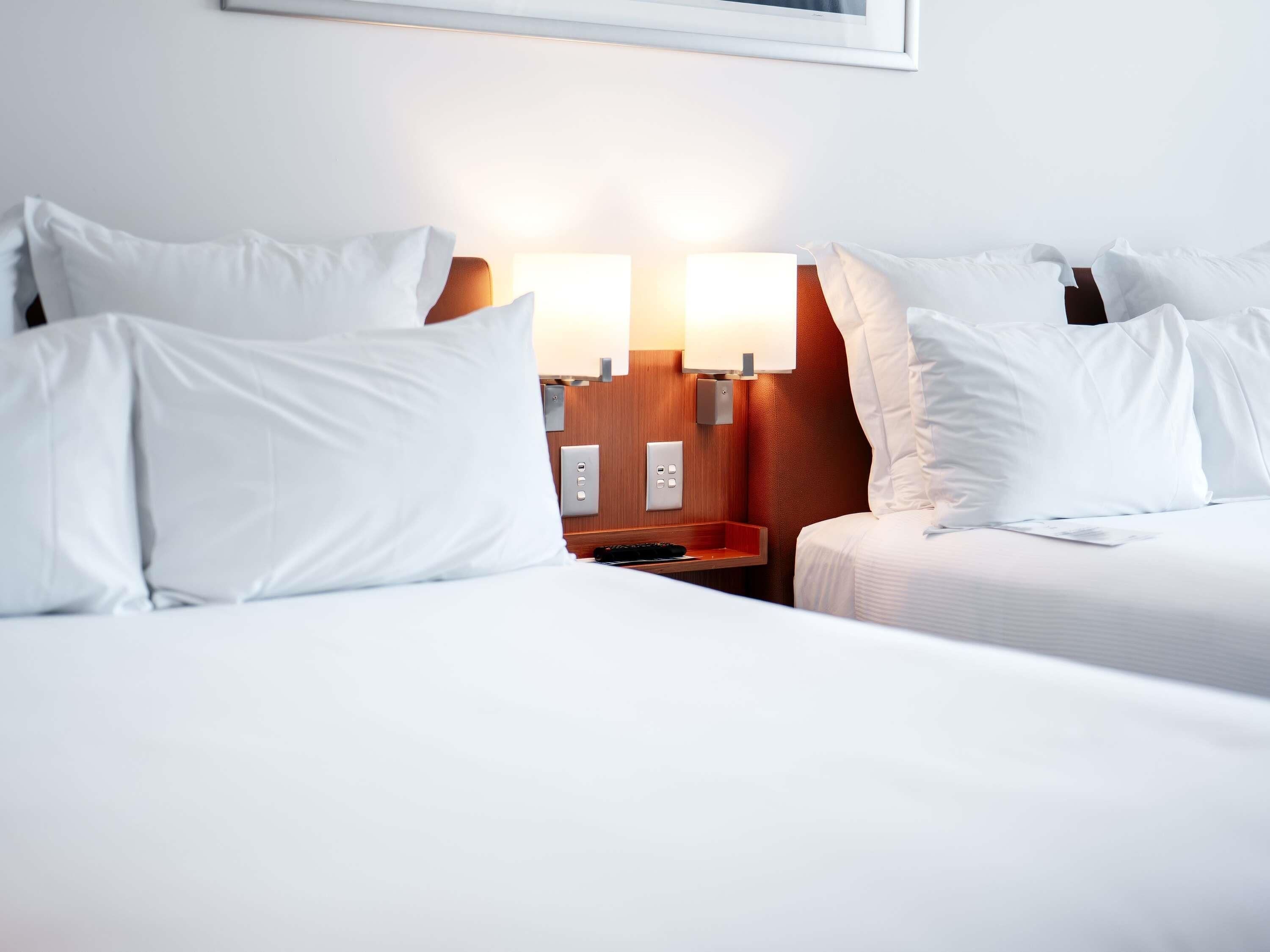 A hotel room with two beds, a nightstand, a lamp, and a wall-mounted light.